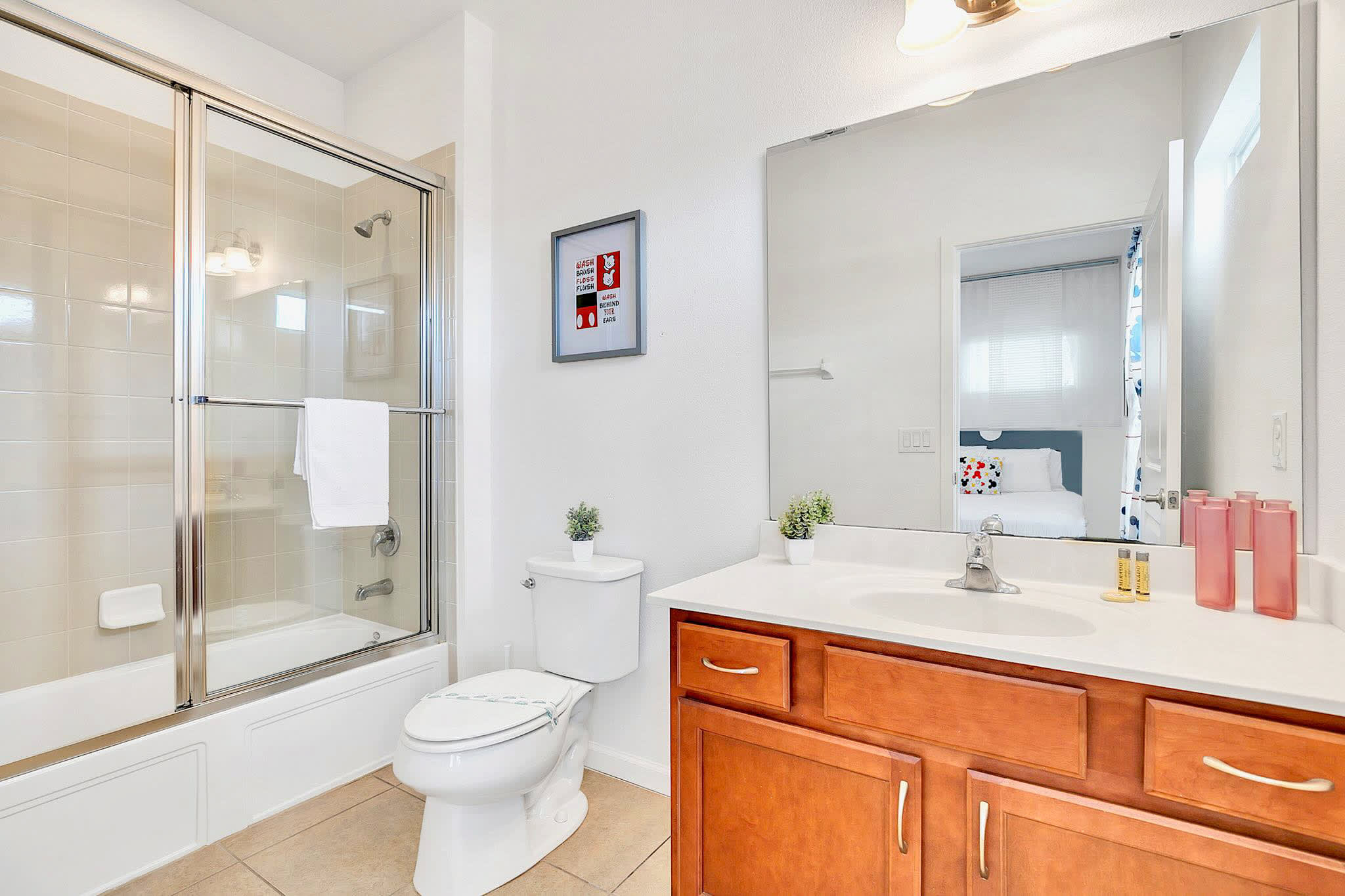 Bright bathroom with a wooden single vanity, white countertop, large mirror, toilet, and a shower/tub combo with a sliding glass door and light tiled walls.
