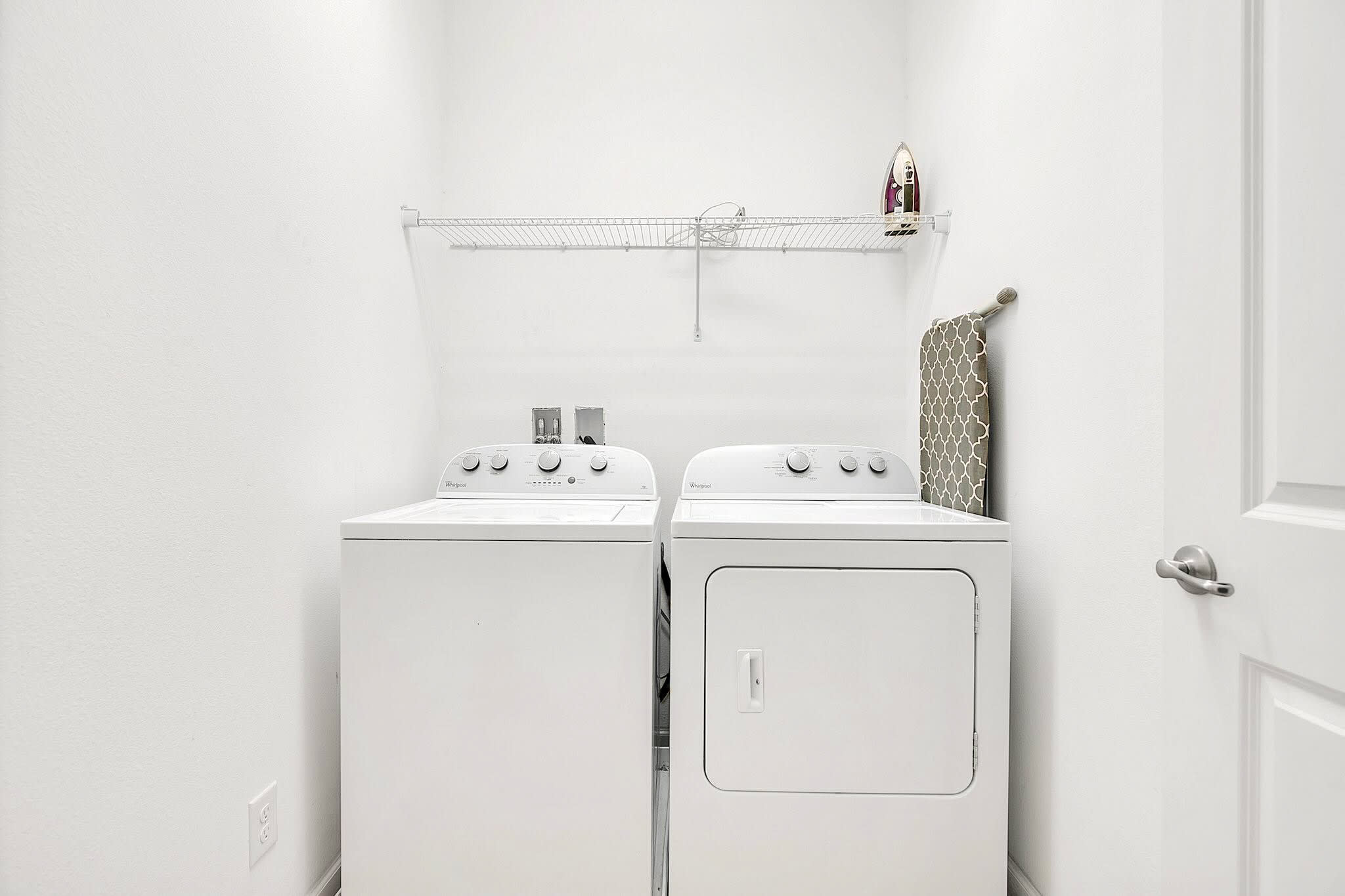 Functional laundry area featuring a white top-loading washing machine and a matching dryer, positioned beneath a wire shelf. An ironing board and iron are stored on the shelf.