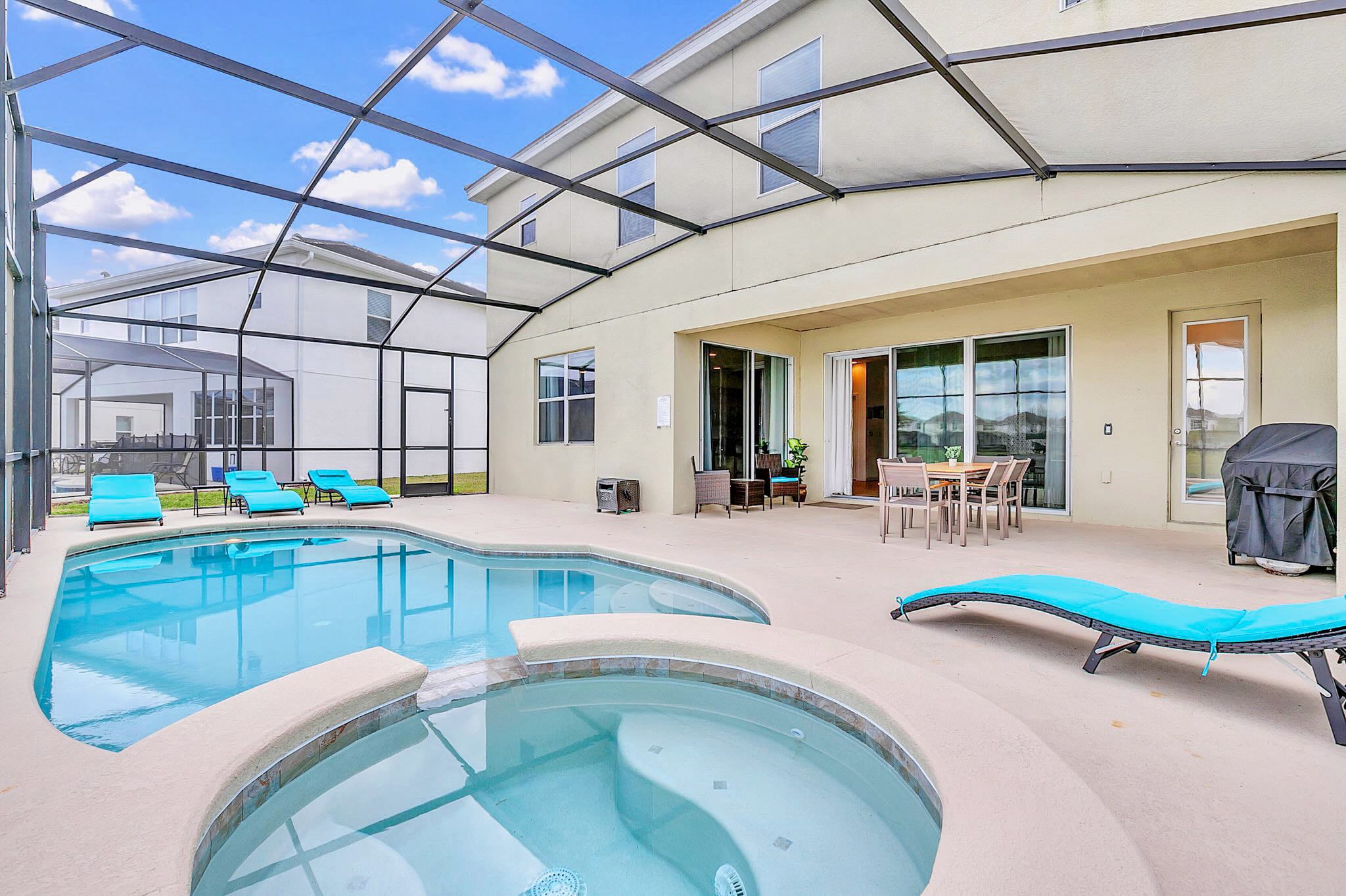Expansive screened-in pool area at the back of a two-story house, featuring a large swimming pool with an integrated hot tub, multiple lounge chairs, and an outdoor dining set, all overlooking a lake.