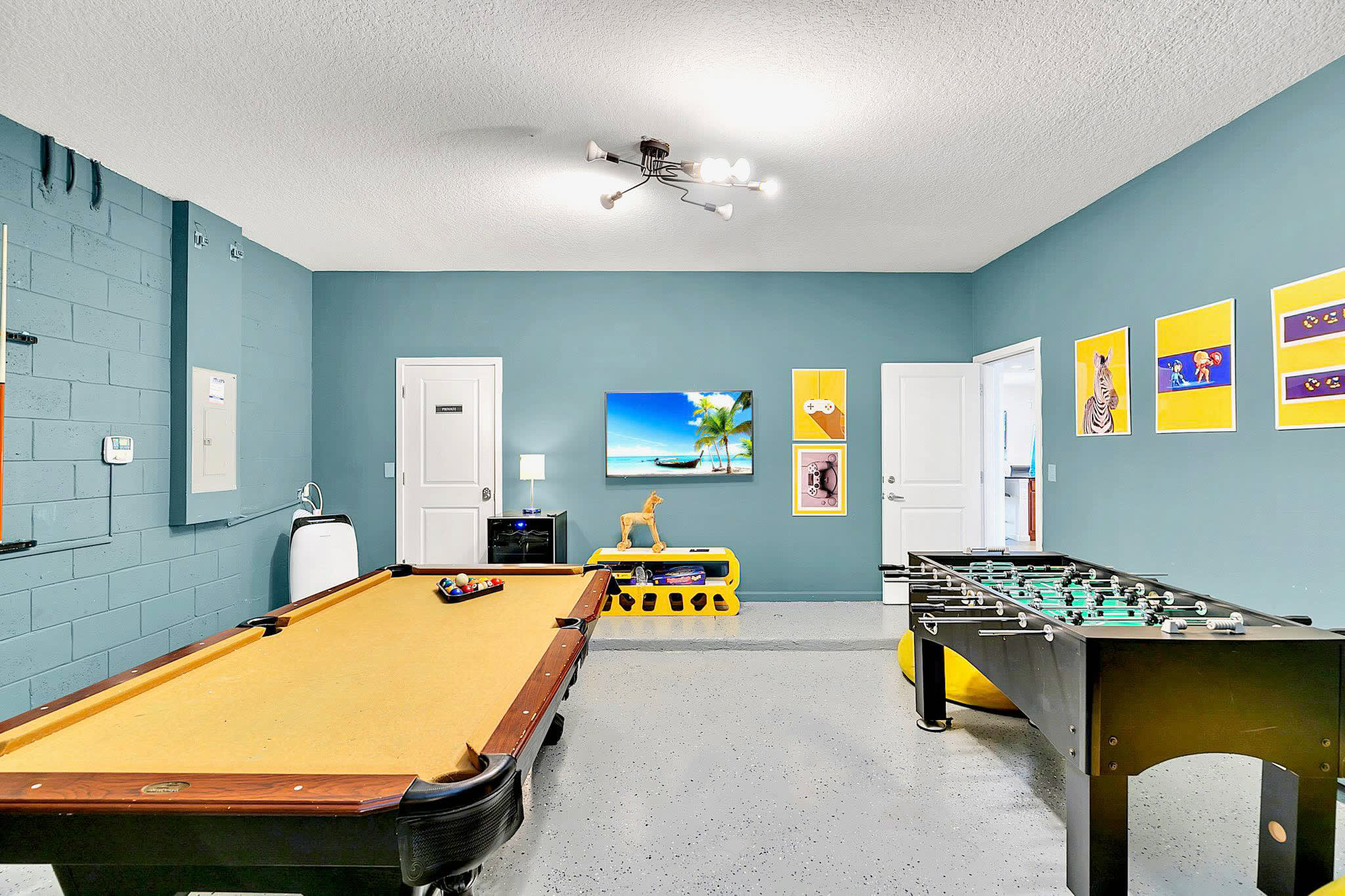 Another angle of the game room, showcasing a large wall-mounted TV, a pool table with yellow felt, and a foosball table, all within a space with teal walls and a light gray speckled floor.