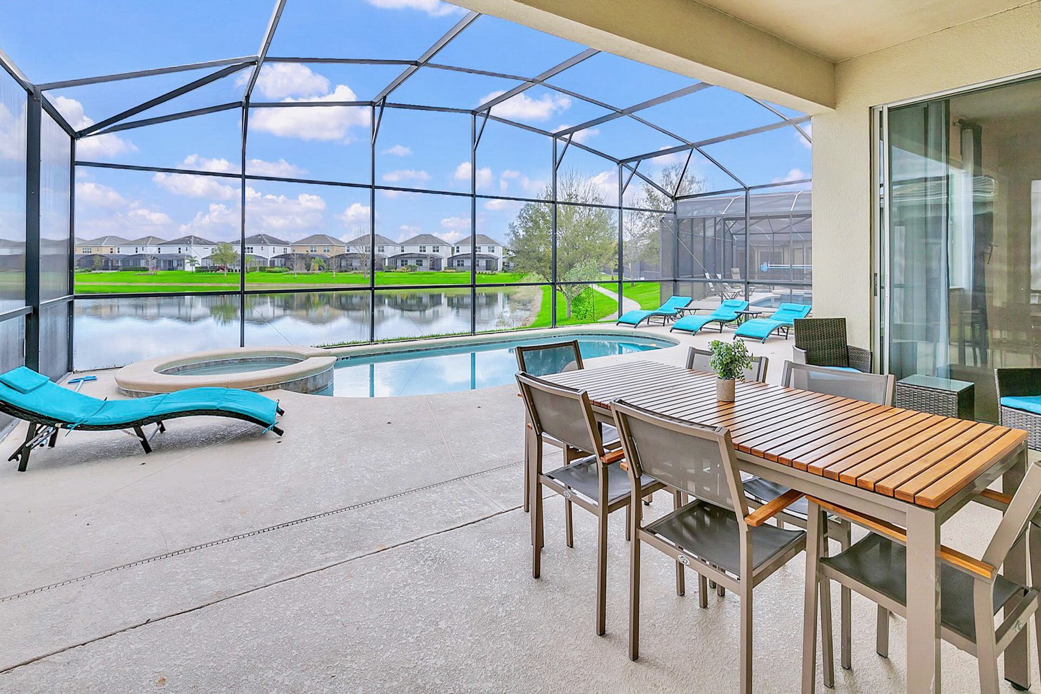 Covered outdoor patio area with a wooden-top dining table and six gray chairs, situated next to a screened-in pool area and overlooking a lake, with sliding glass doors leading into the house.