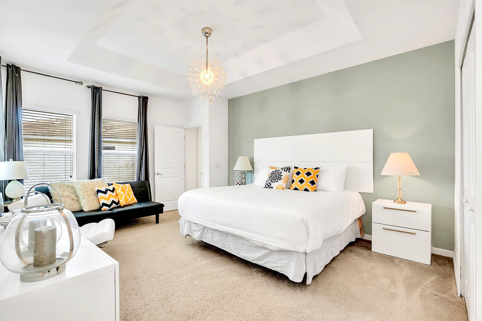 Spacious master bedroom featuring a white king-sized bed with a large headboard, a light green accent wall, a black futon, and a tray ceiling with a modern chandelier.