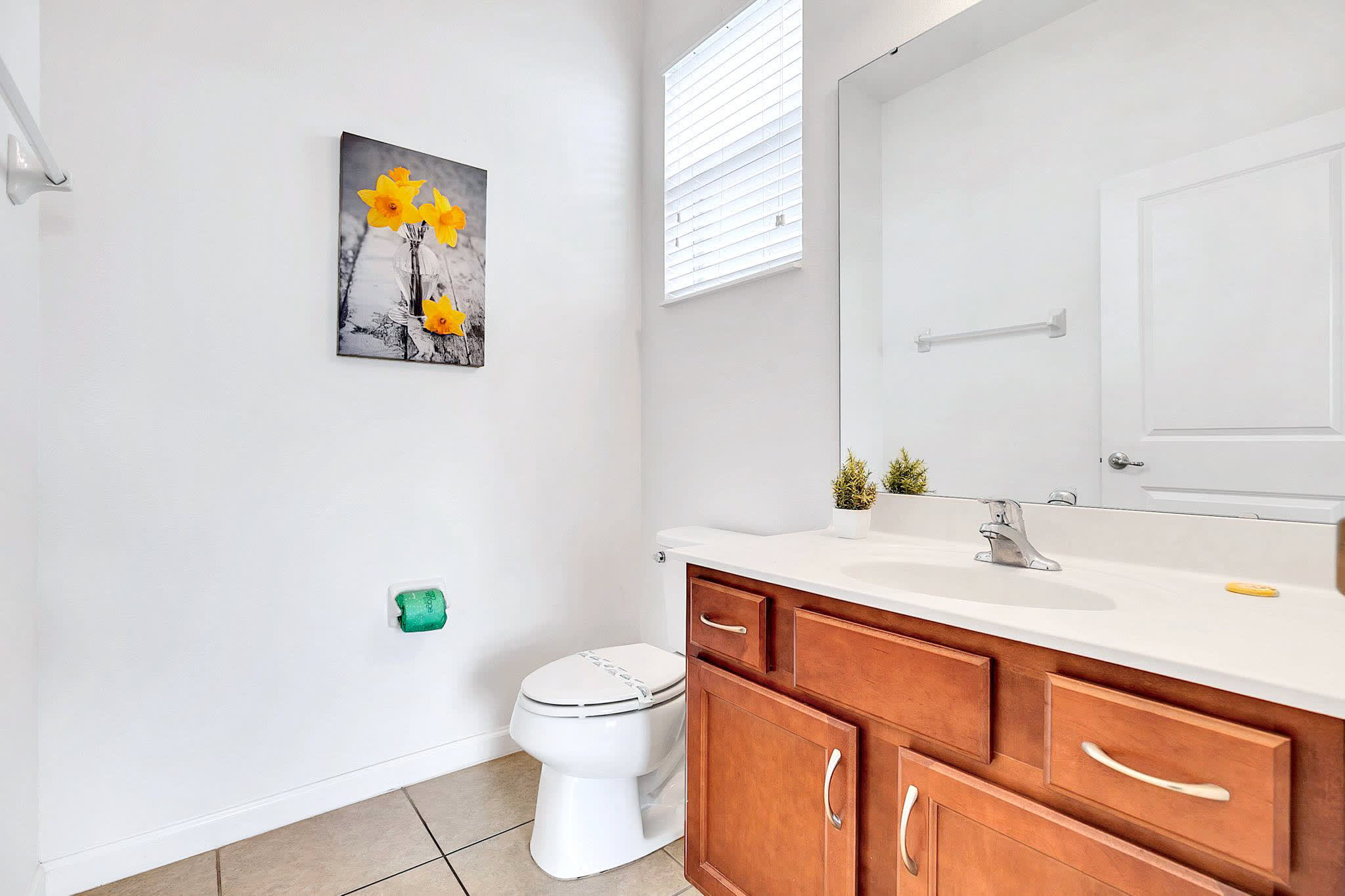 Clean white bathroom with a toilet, a wooden vanity with a white countertop and sink, and a large mirror. A small window with blinds is visible above the toilet, and a yellow floral art piece adorns the wall.