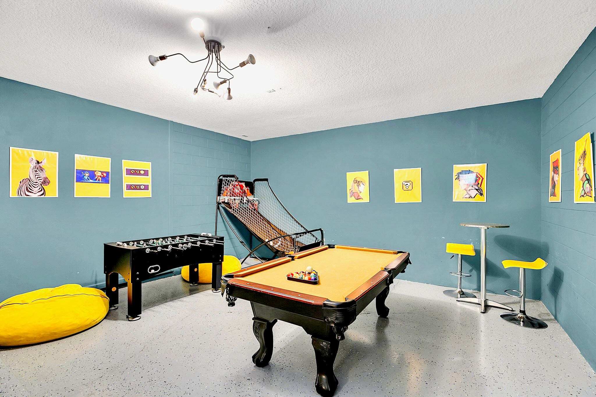 Vibrant game room with teal walls and a speckled gray floor, featuring a pool table, foosball table, arcade basketball game, and a high-top table with yellow stools, decorated with colorful pop art.