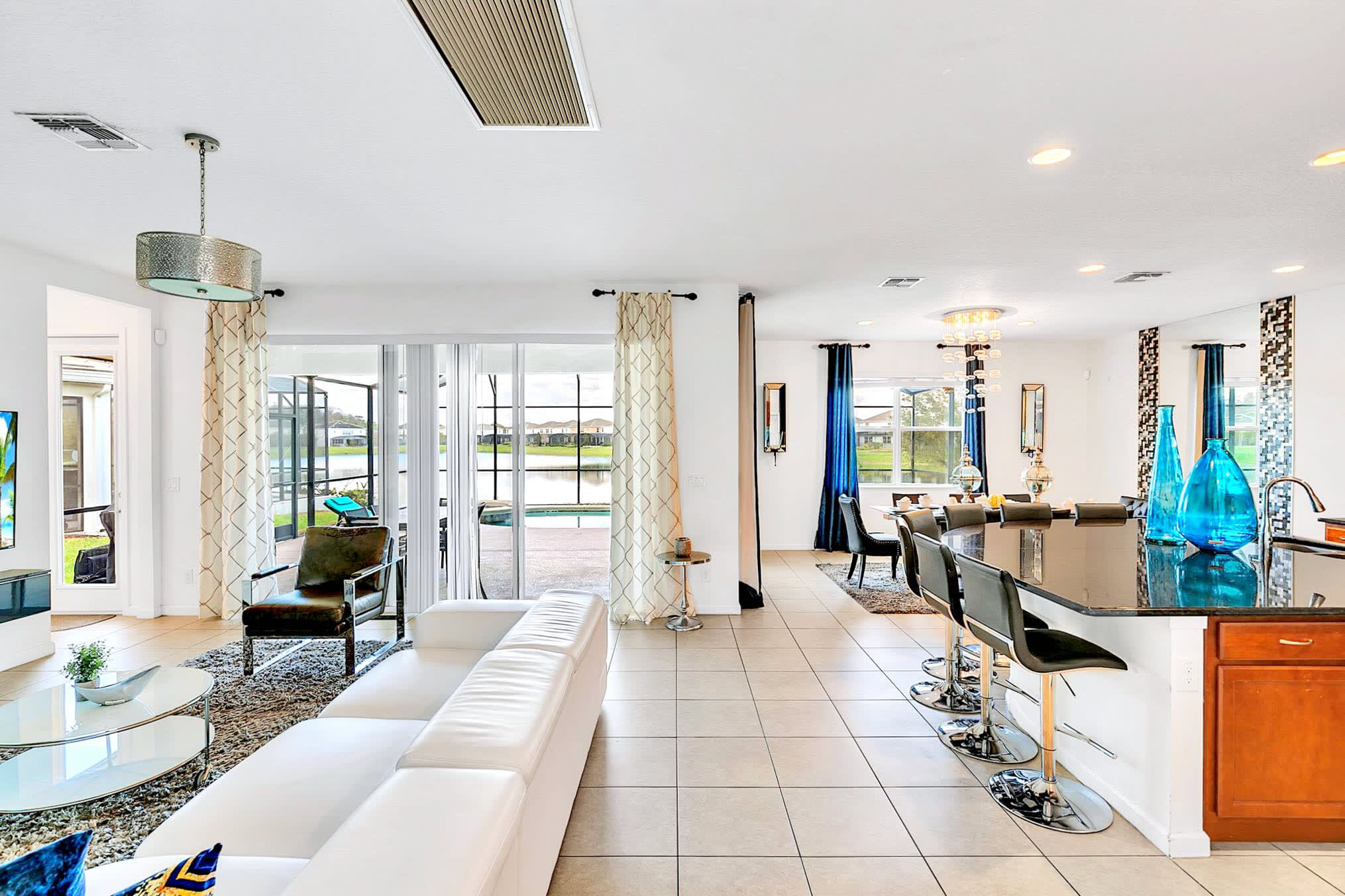 Open-concept living space connecting the living room with a white sectional to a kitchen island with bar seating, and a dining area beyond, all with light tile flooring and views to the outside.