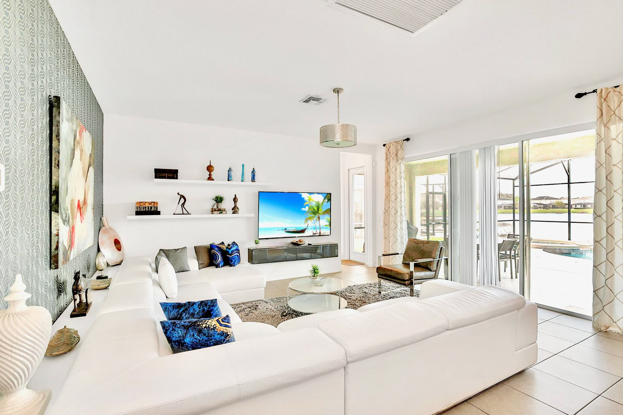 Bright living room with a white sectional sofa and a large TV, offering views of an outdoor pool and lake through expansive sliding glass doors, complemented by light tile flooring.