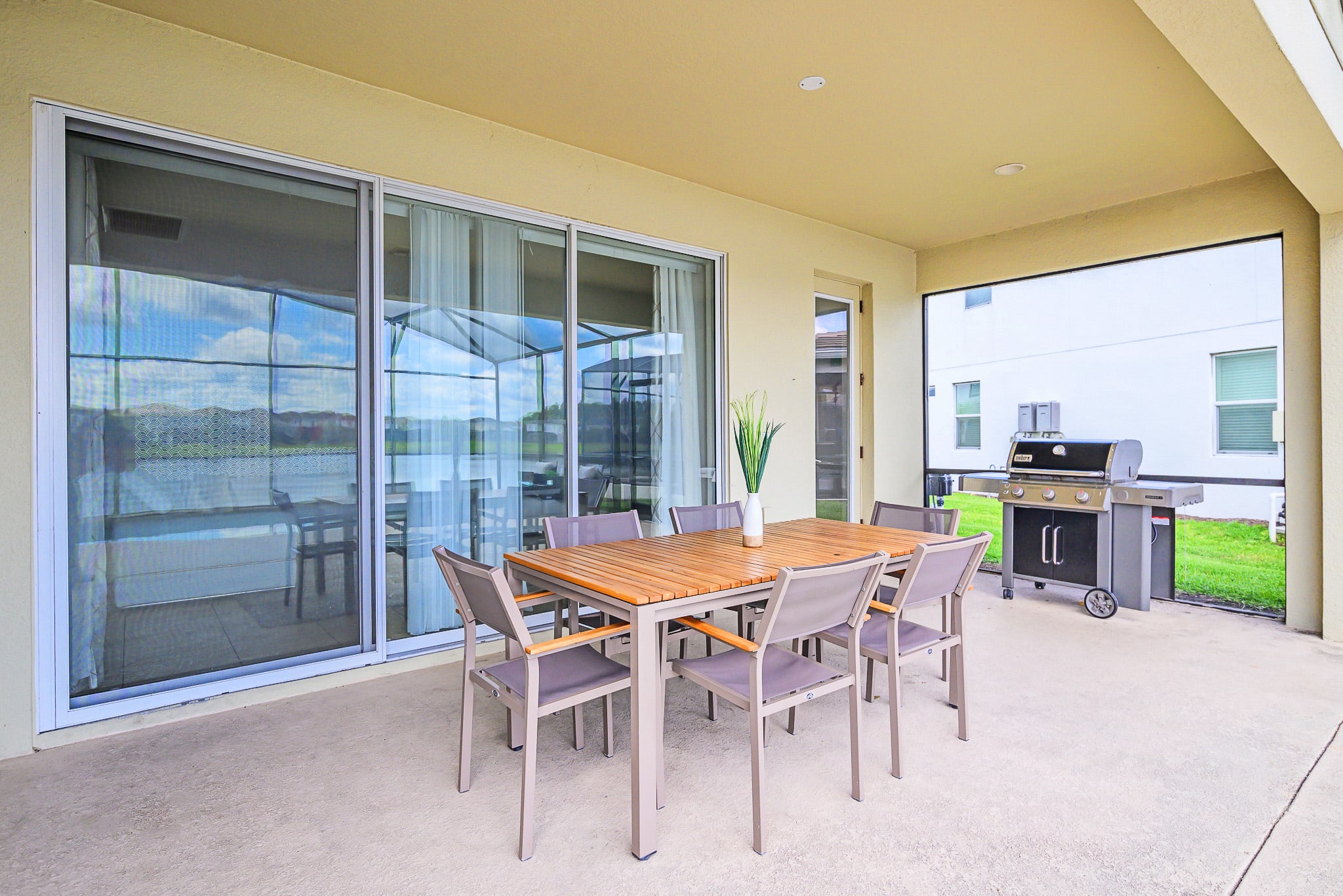 Covered outdoor patio featuring a wooden-top dining table with gray chairs and a stainless steel BBQ grill, with large sliding glass doors opening to the interior of the house and a view of the screened pool area.