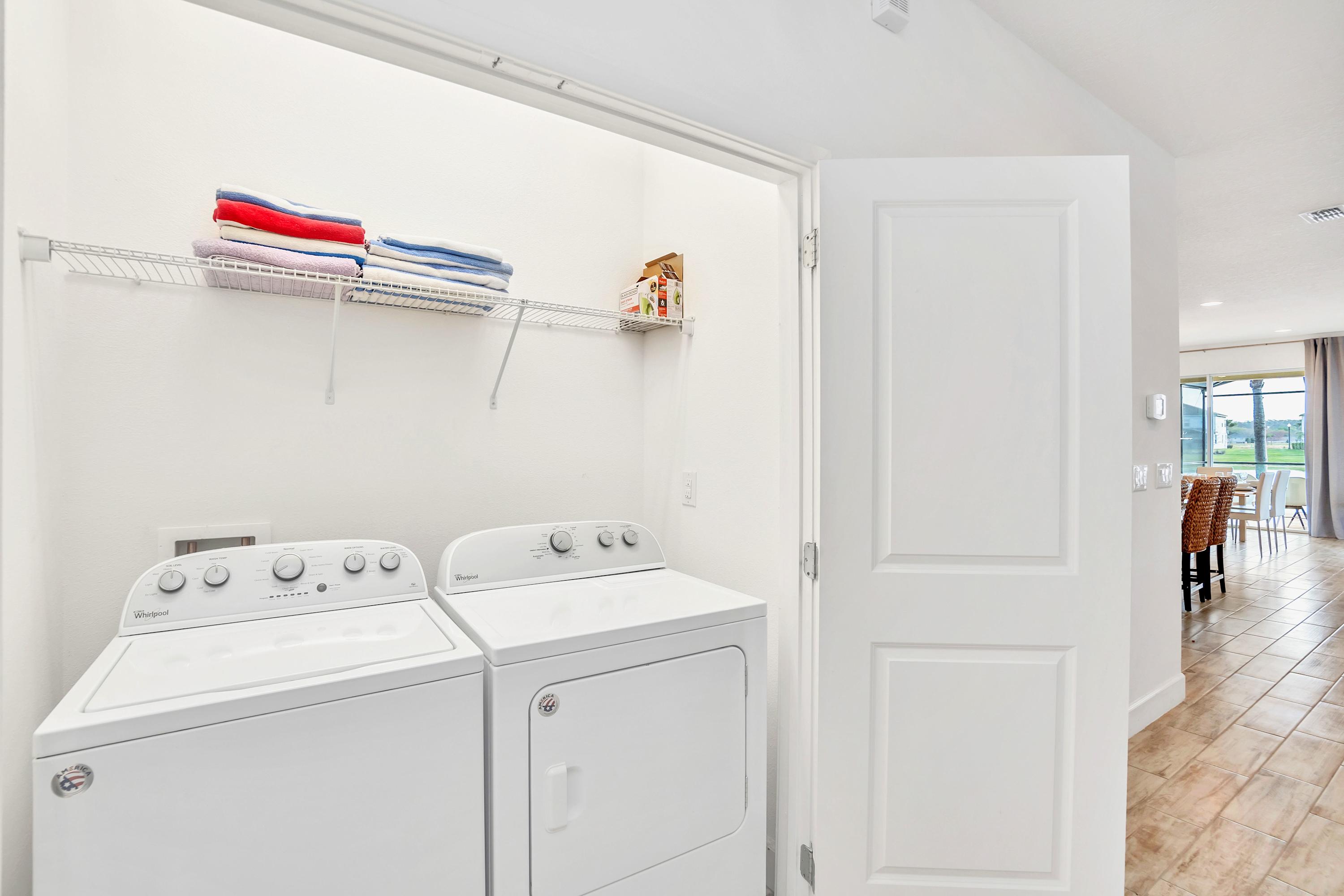 A bright laundry closet equipped with a white top-loading washing machine and a matching dryer, stacked with neatly folded towels on a wire shelf above. The space is clean and functional, with white walls and a view into an adjacent room through an open door.