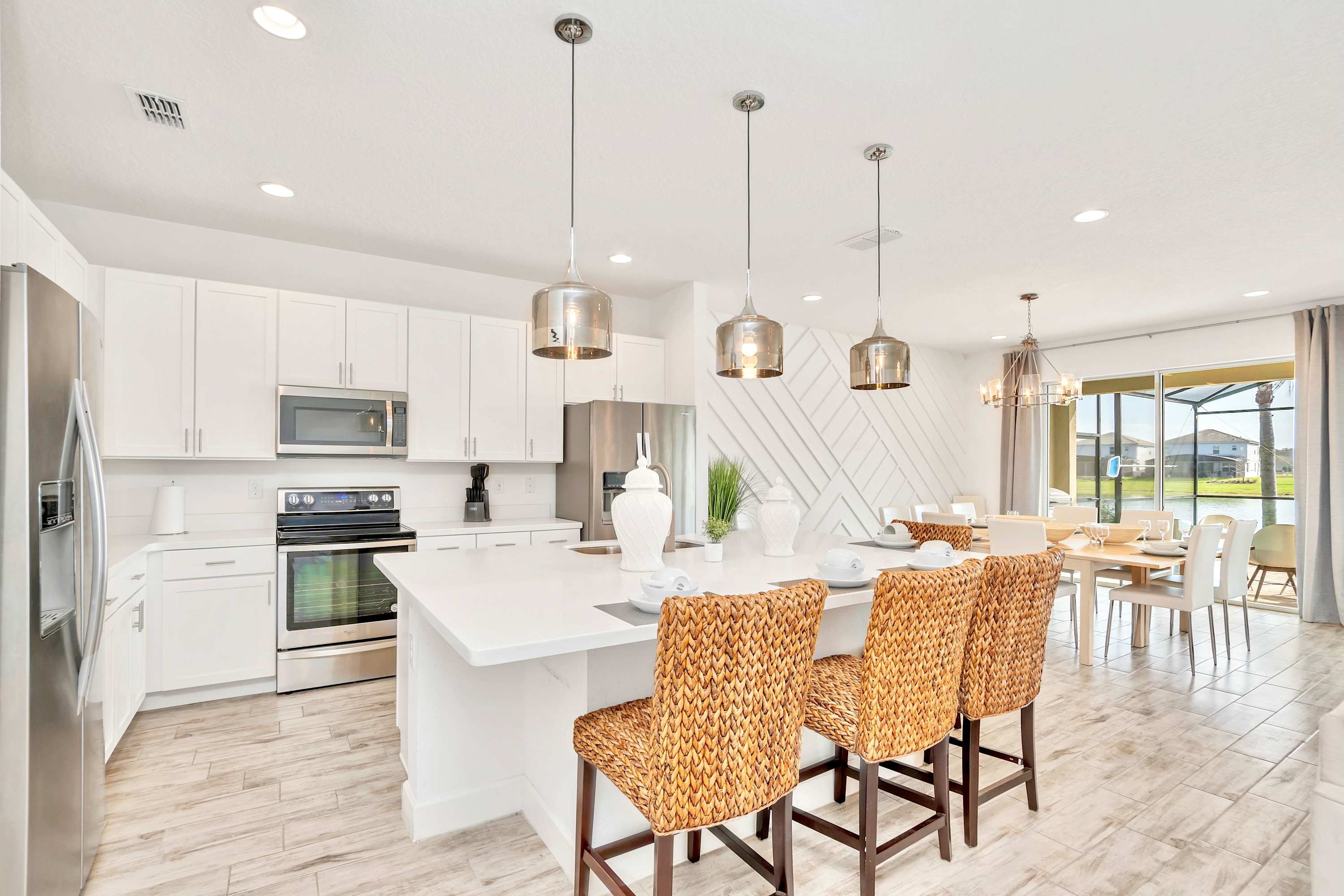 A modern kitchen boasts white shaker cabinets, white quartz countertops, and stainless steel appliances, including an oven and refrigerator. A large island with three wicker bar stools and contemporary pendant lights serves as a central gathering spot, all on light wood-look tile flooring.