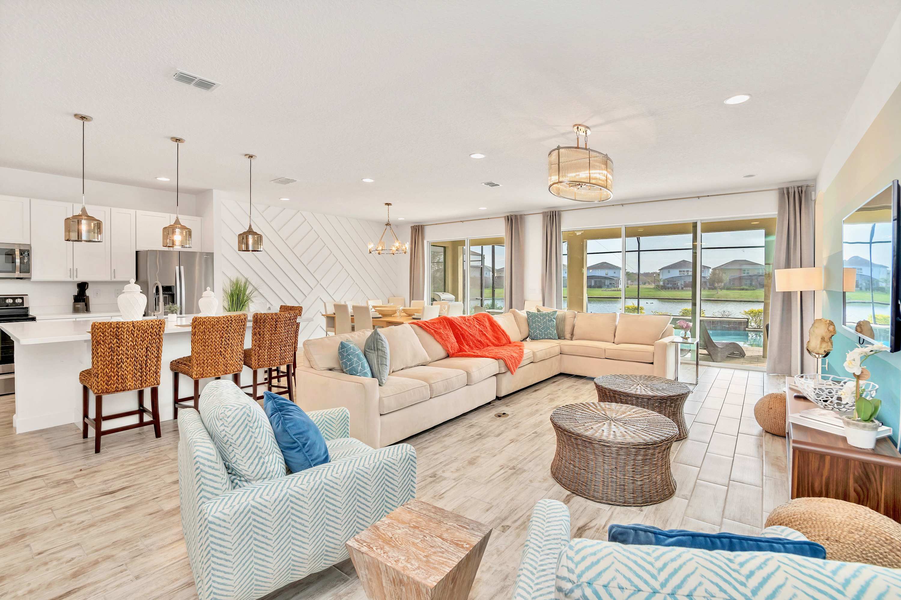 An expansive open-concept living area features a large L-shaped sectional sofa with colorful throw pillows, facing a wall-mounted TV. The space seamlessly connects to a modern kitchen with a breakfast bar and a dining area, all with light wood-look flooring and large sliding glass doors opening to the pool.