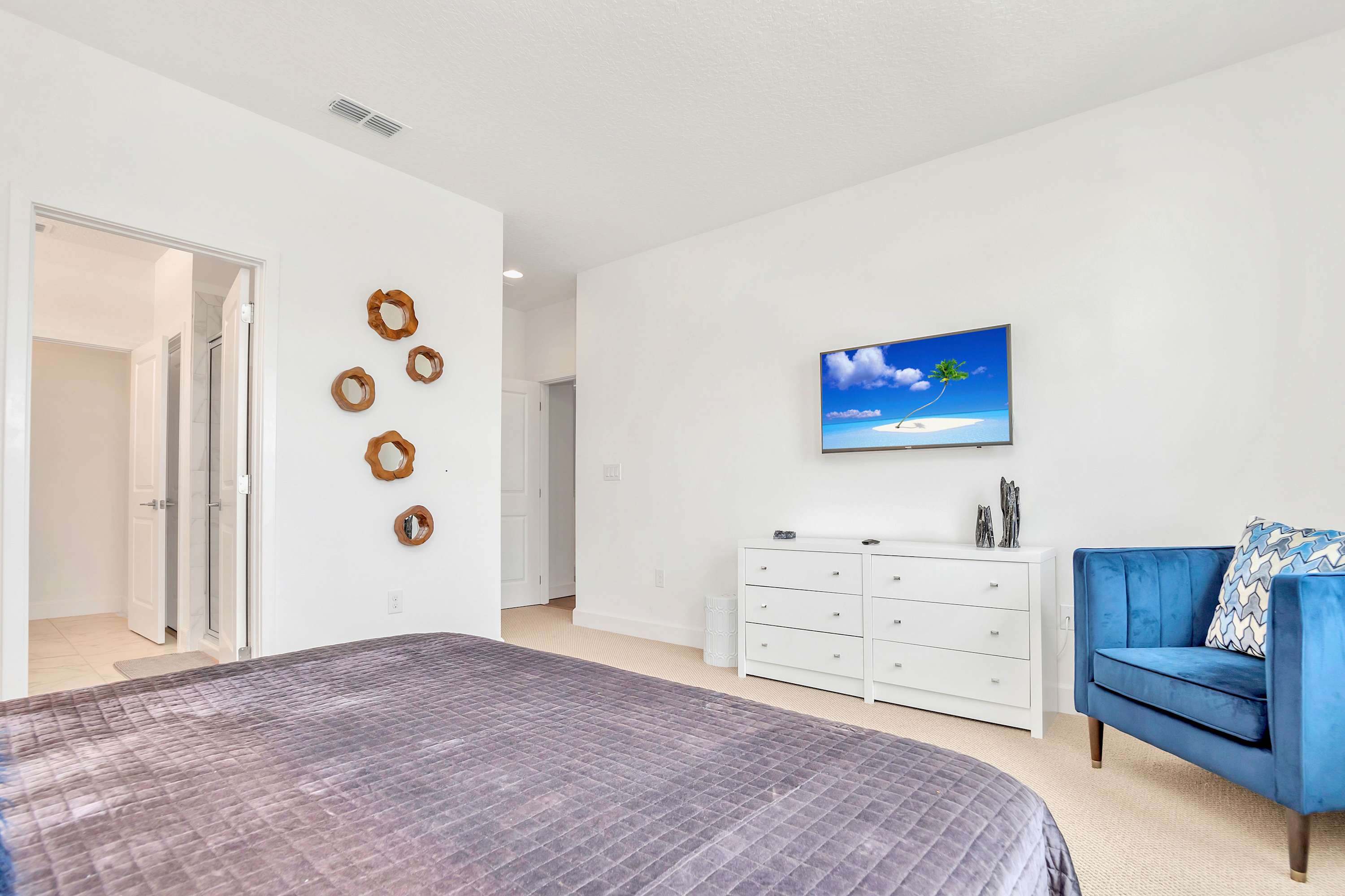 This bedroom features a king-size bed with purple bedding, a blue velvet armchair, and a white dresser with a wall-mounted TV. The room is carpeted, and a doorway leads to an en-suite bathroom or closet.