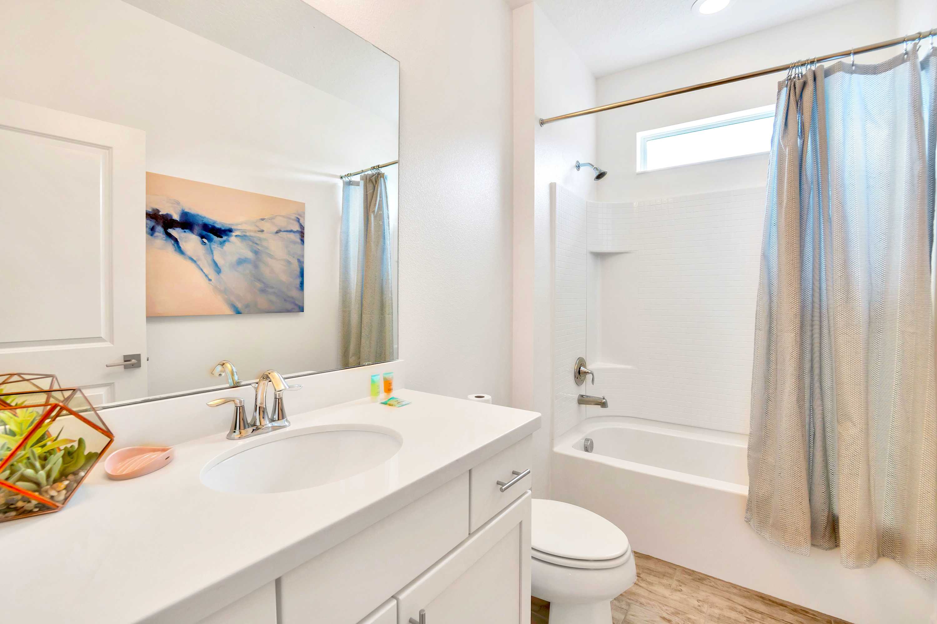 Bright white bathroom with a single vanity, large mirror, toilet, and a shower-tub combo with a light gray shower curtain. Wood-look tile flooring adds warmth to the space.