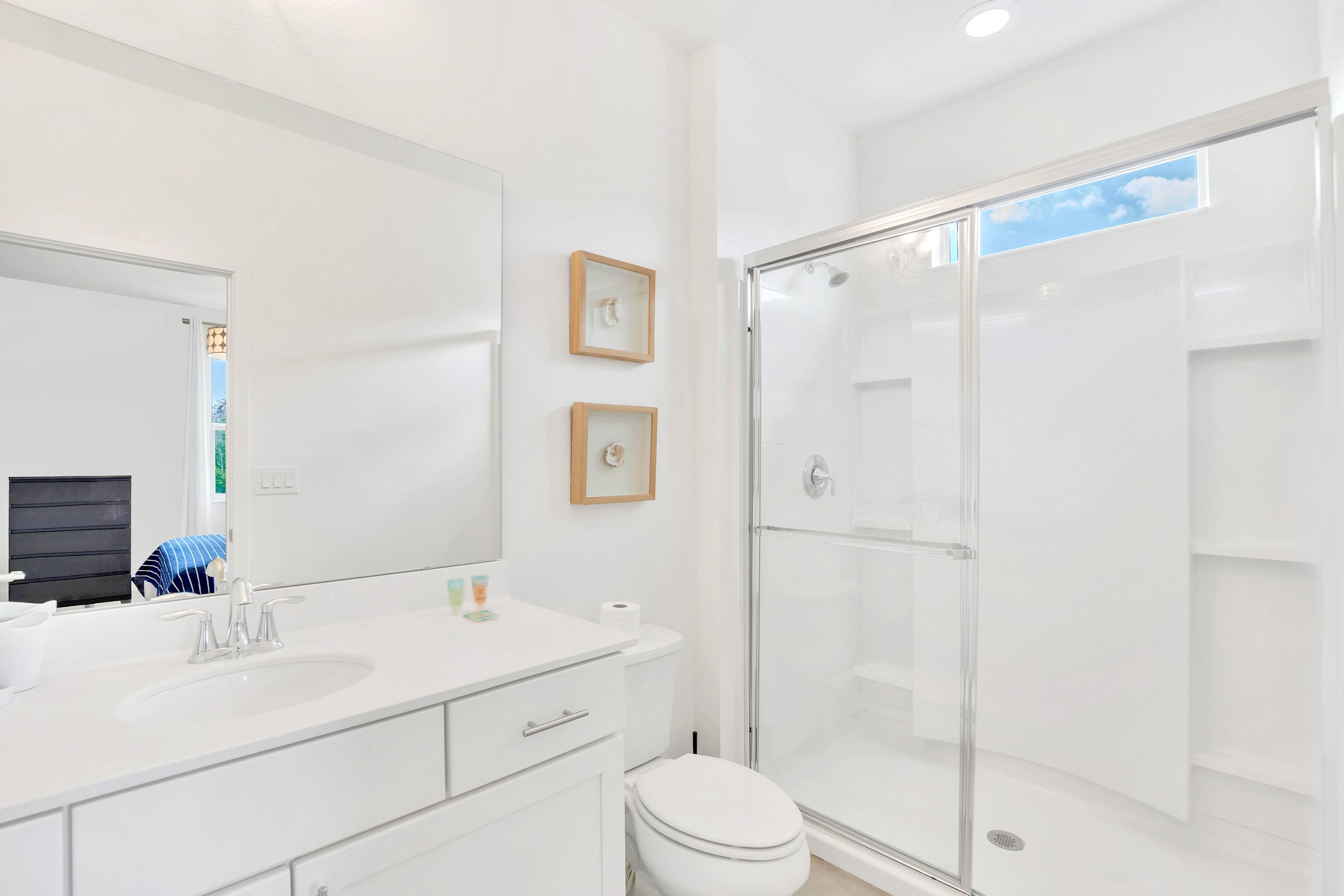 Modern white bathroom with a single vanity, large mirror, toilet, and a walk-in shower with a clear glass enclosure. Shell-themed framed artwork decorates the wall.