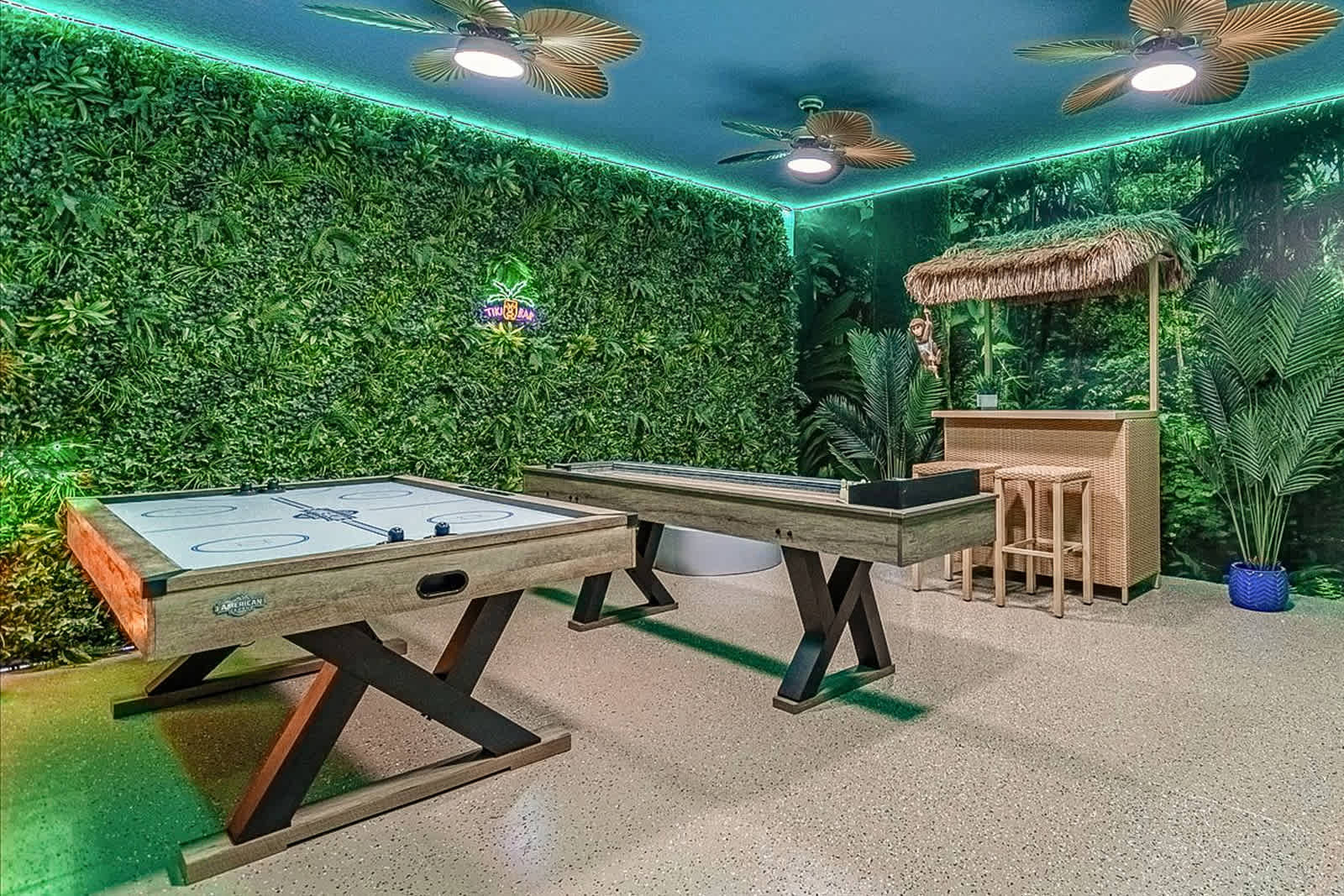 A vibrant jungle-themed game room features a green artificial plant wall, a shuffleboard table, an air hockey table, and a small tiki bar with stools. The room is illuminated by green LED strip lights and ceiling fans, with a light-colored speckled floor.