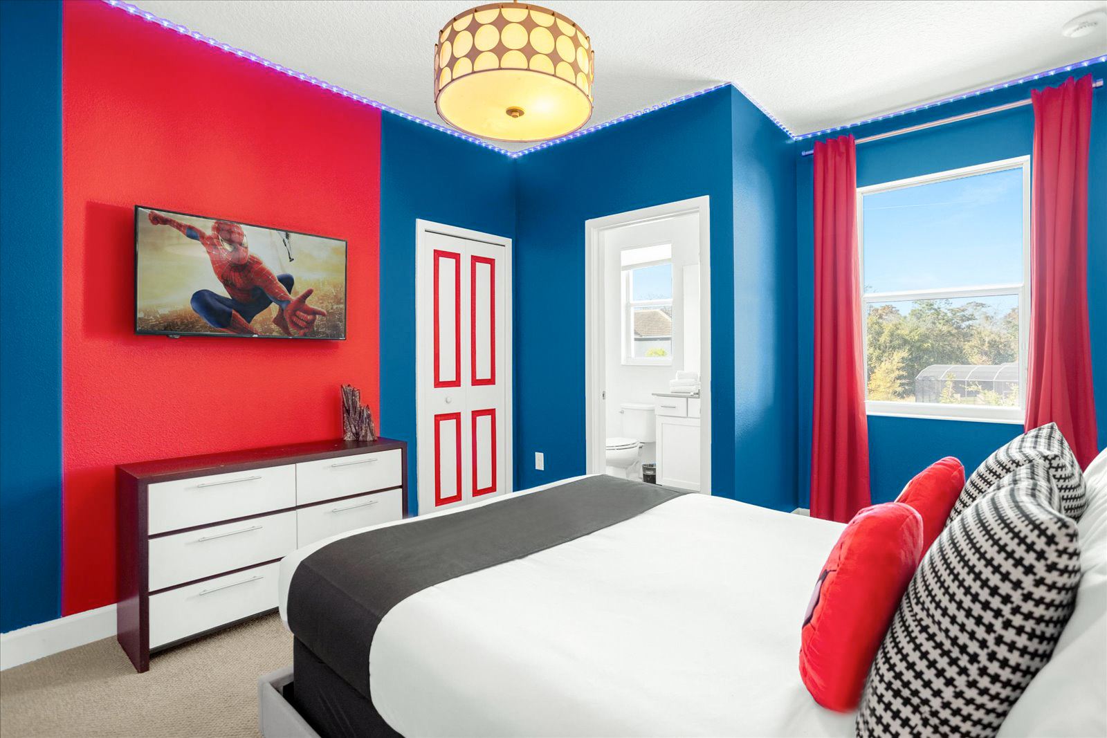 Vibrant themed bedroom with a red accent wall and blue walls, featuring a wall-mounted TV showing Spiderman. A white dresser with dark drawers sits below the TV, and red curtains frame a window.