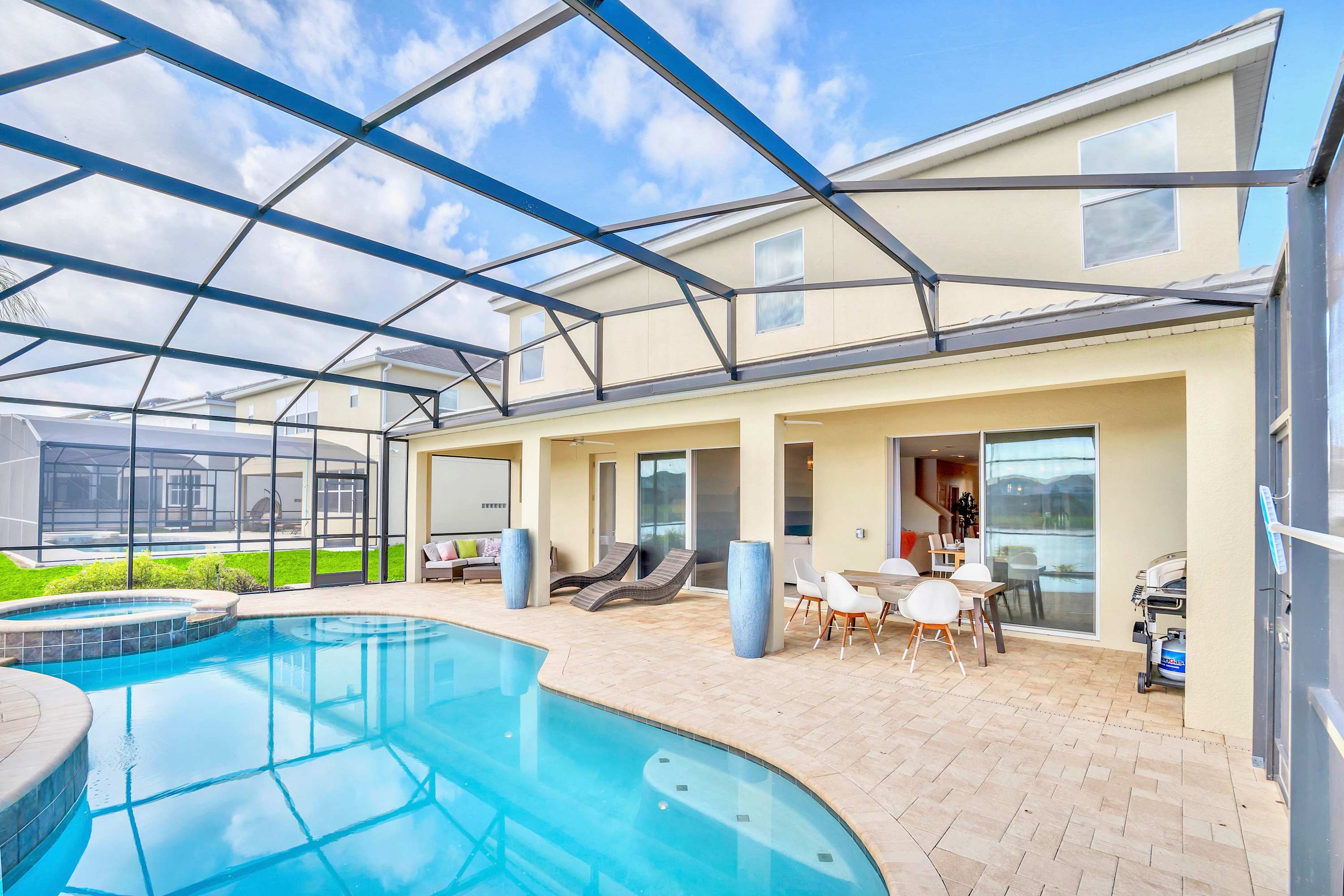 A spacious screened-in outdoor pool area features a light blue swimming pool with an integrated hot tub, surrounded by a paved patio. The area is furnished with lounge chairs, an outdoor dining set, and a BBQ grill, all overlooking a serene lake and the back of the house.