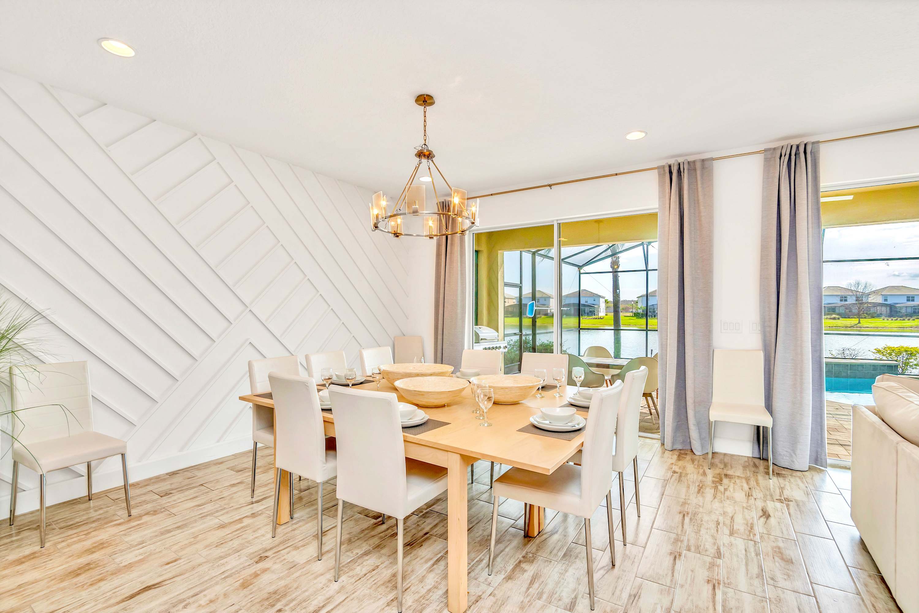 A bright dining room features a large rectangular wooden table with eight white chairs, set against a striking white geometric accent wall. Sliding glass doors open to a screened patio with a pool and a lake view, all on light wood-look tile flooring.