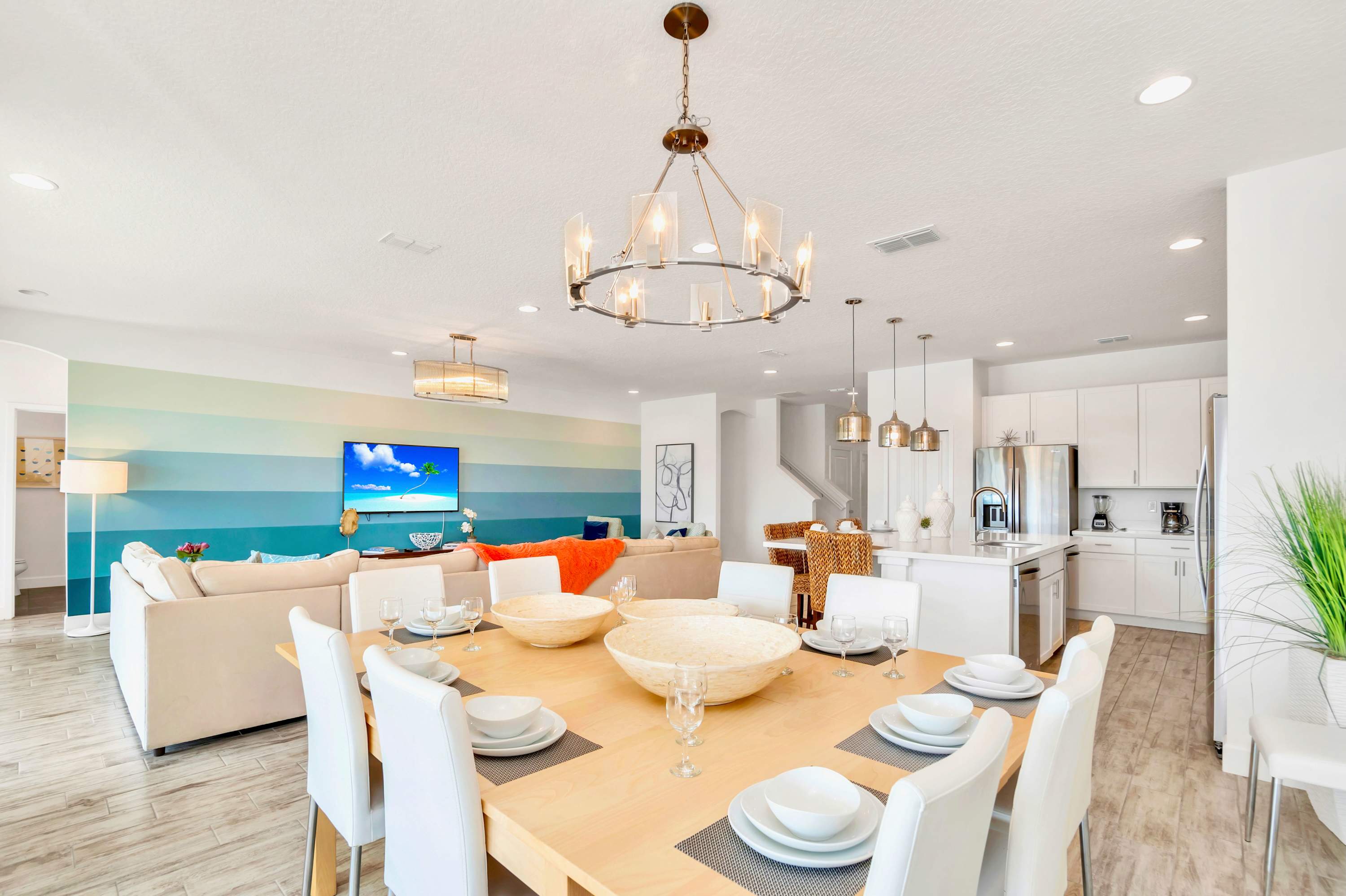 A spacious dining room features a large light wood rectangular table with eight white chairs, illuminated by a modern chandelier. It's part of an open-concept layout, with a living room and kitchen visible in the background, all on light wood-look tile flooring.