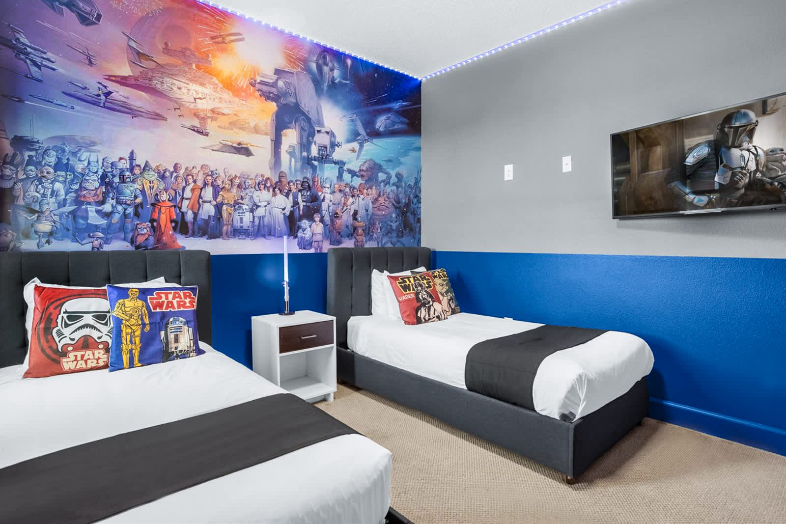 Star Wars-themed bedroom with a vibrant wall mural and a wall-mounted TV displaying "The Mandalorian." Two twin beds with themed pillows are visible, separated by a nightstand with a light saber lamp.