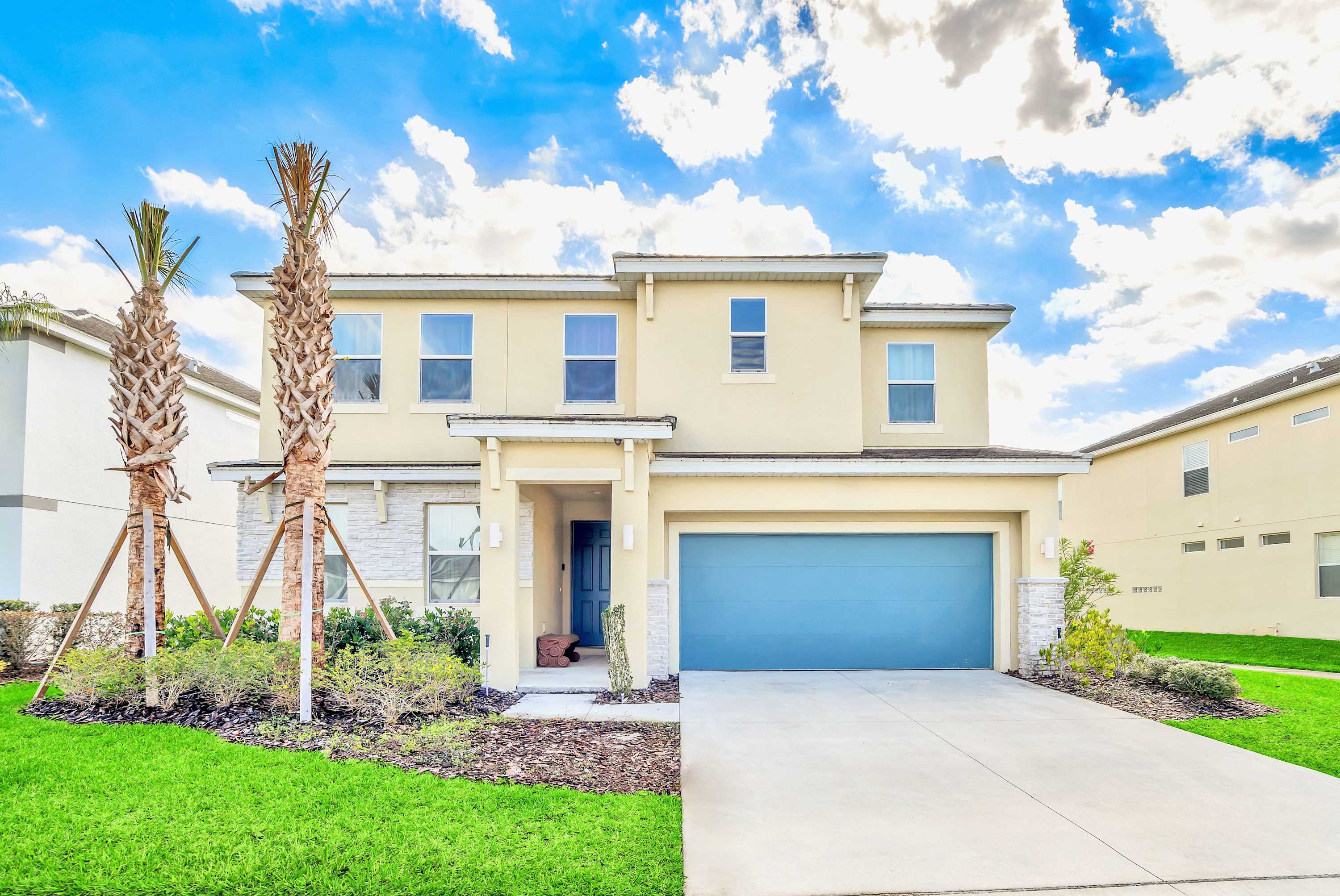 A two-story, light yellow stucco house with stone accents features a bright blue garage door and a matching blue front door. Two tall, slender palm trees flank the entrance, set against a vibrant green lawn and a clear blue sky with fluffy clouds.