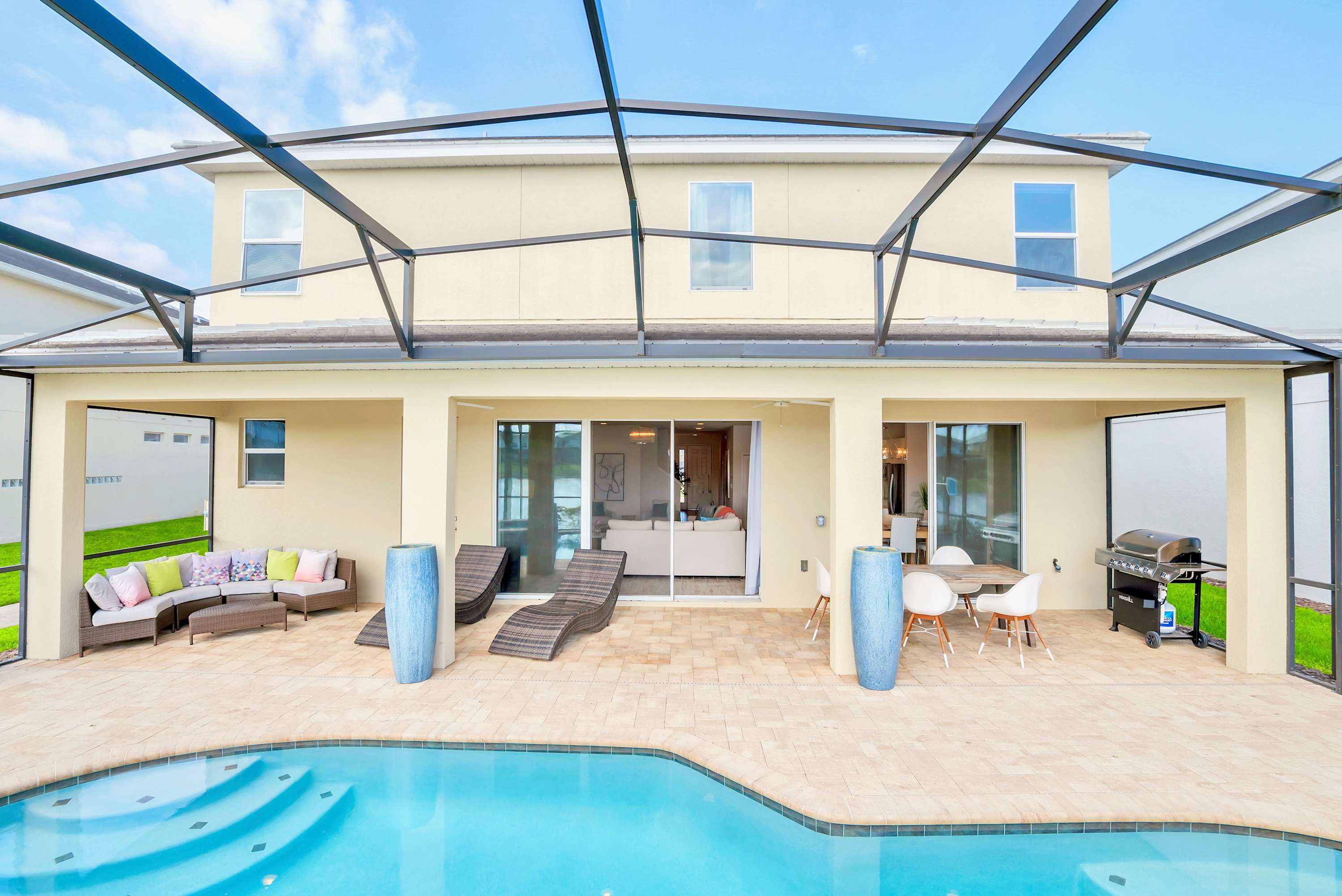 The back exterior of a large, two-story house with light stucco siding features a screened-in patio with a private pool and hot tub. The patio is well-furnished with outdoor seating, lounge chairs, and a dining table, all overlooking a tranquil lake.