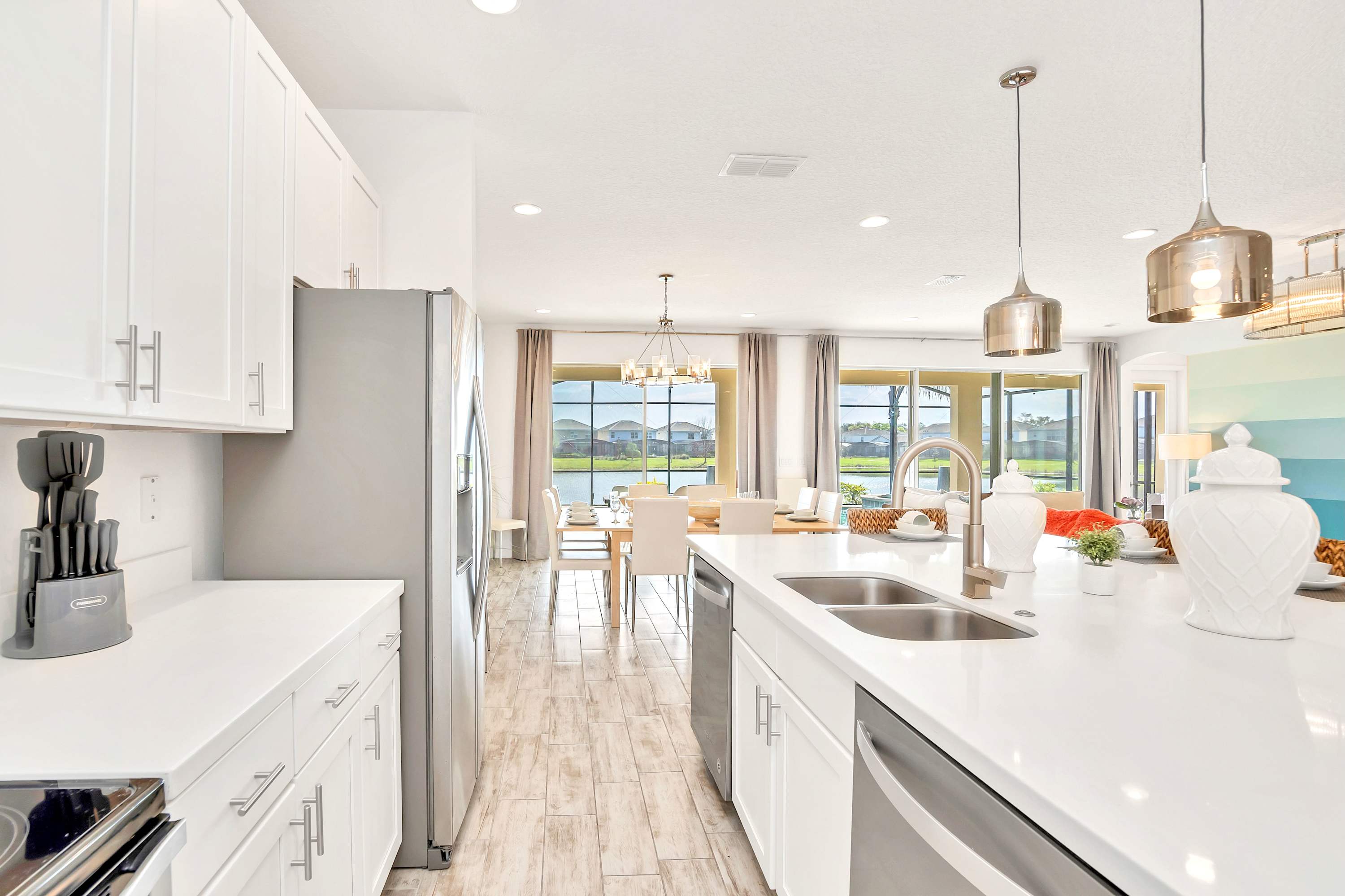A bright kitchen features white cabinets and quartz countertops with a double sink and stainless steel faucet, offering a clear view into the dining area and out to the lake and pool through large sliding glass doors. Stainless steel appliances are integrated throughout.