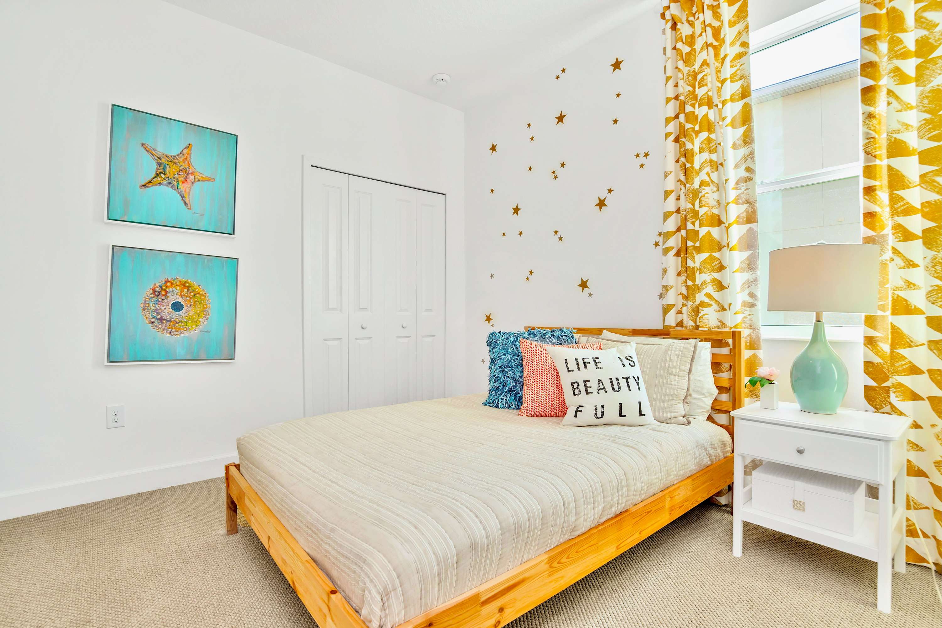 Bright bedroom with a full-size wooden bed frame, white walls adorned with star decals, and vibrant yellow and white patterned curtains. A white nightstand with a lamp is beside the bed.