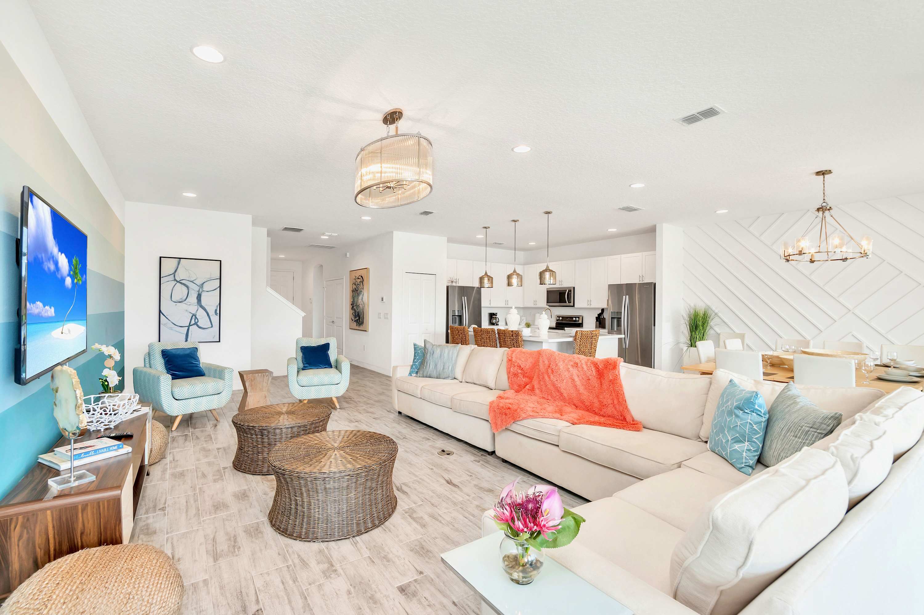 A bright, open-concept living room features a large L-shaped sectional sofa with colorful throws, two light blue armchairs, and wicker coffee tables on light wood-look tile flooring. A large TV is mounted on a wall with a blue and white striped accent, and a modern chandelier hangs from the ceiling.