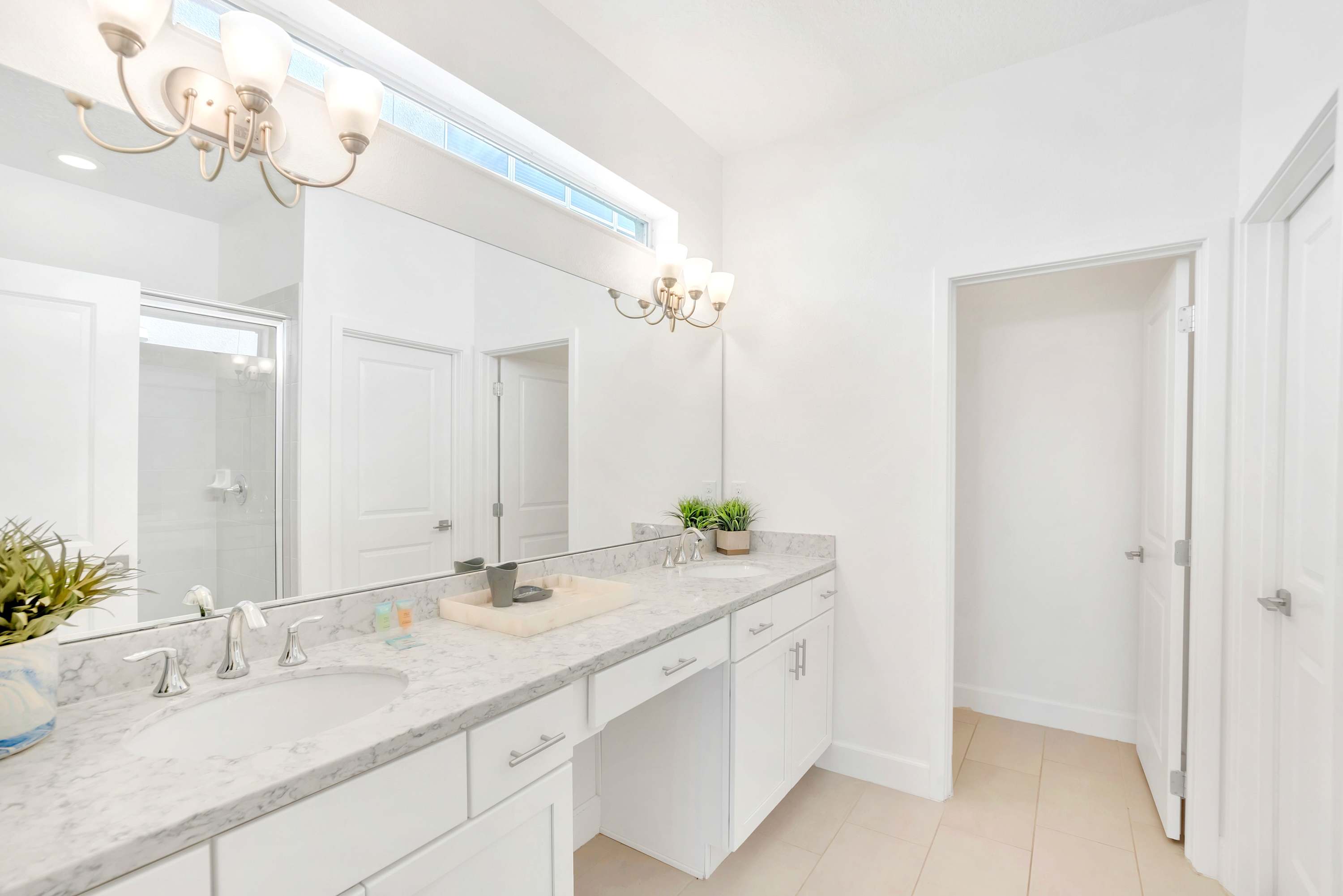 Bright white bathroom with a double vanity featuring a light granite countertop, two sinks, and chrome faucets. A large mirror spans the wall, illuminated by two multi-bulb light fixtures.