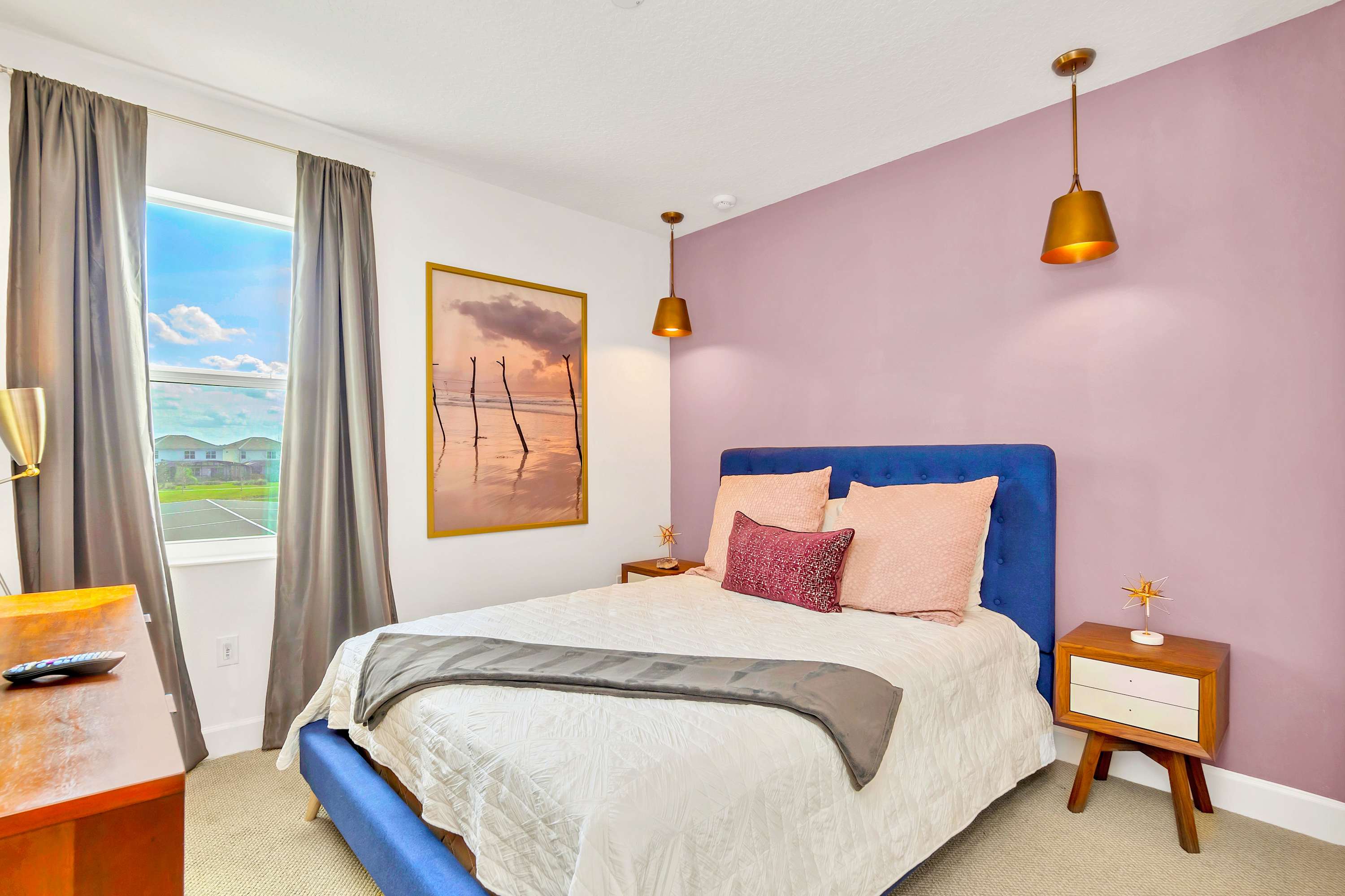 A vibrant bedroom featuring a blue upholstered bed with white and pink bedding, set against a lavender accent wall, and illuminated by two brass pendant lights. A large window with grey curtains offers a view of houses and sky, while a framed beach art piece hangs above the bed.