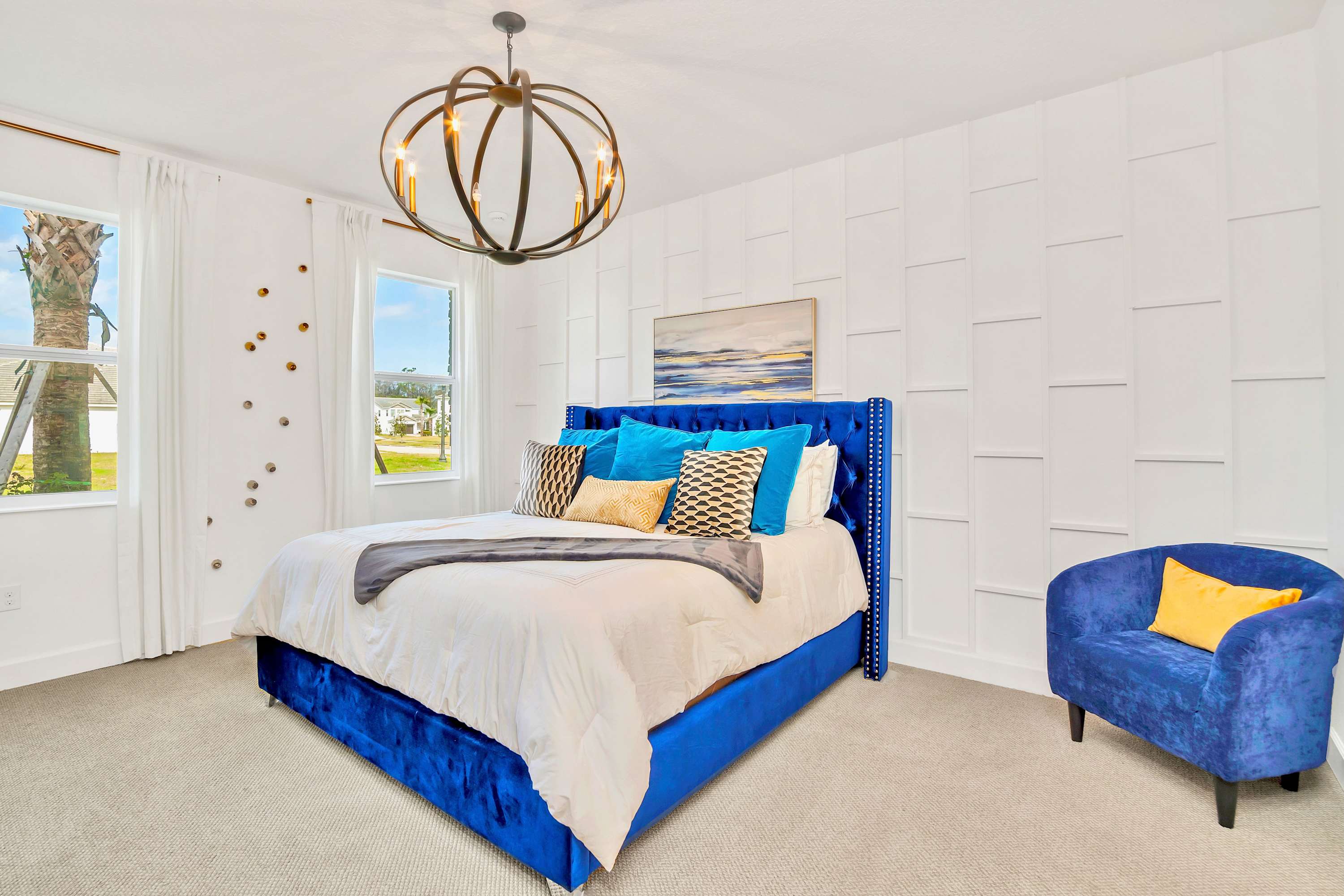 White-walled bedroom featuring a royal blue velvet tufted bed with white bedding, a matching blue armchair, and a modern spherical chandelier. The room has a textured accent wall and a large window.