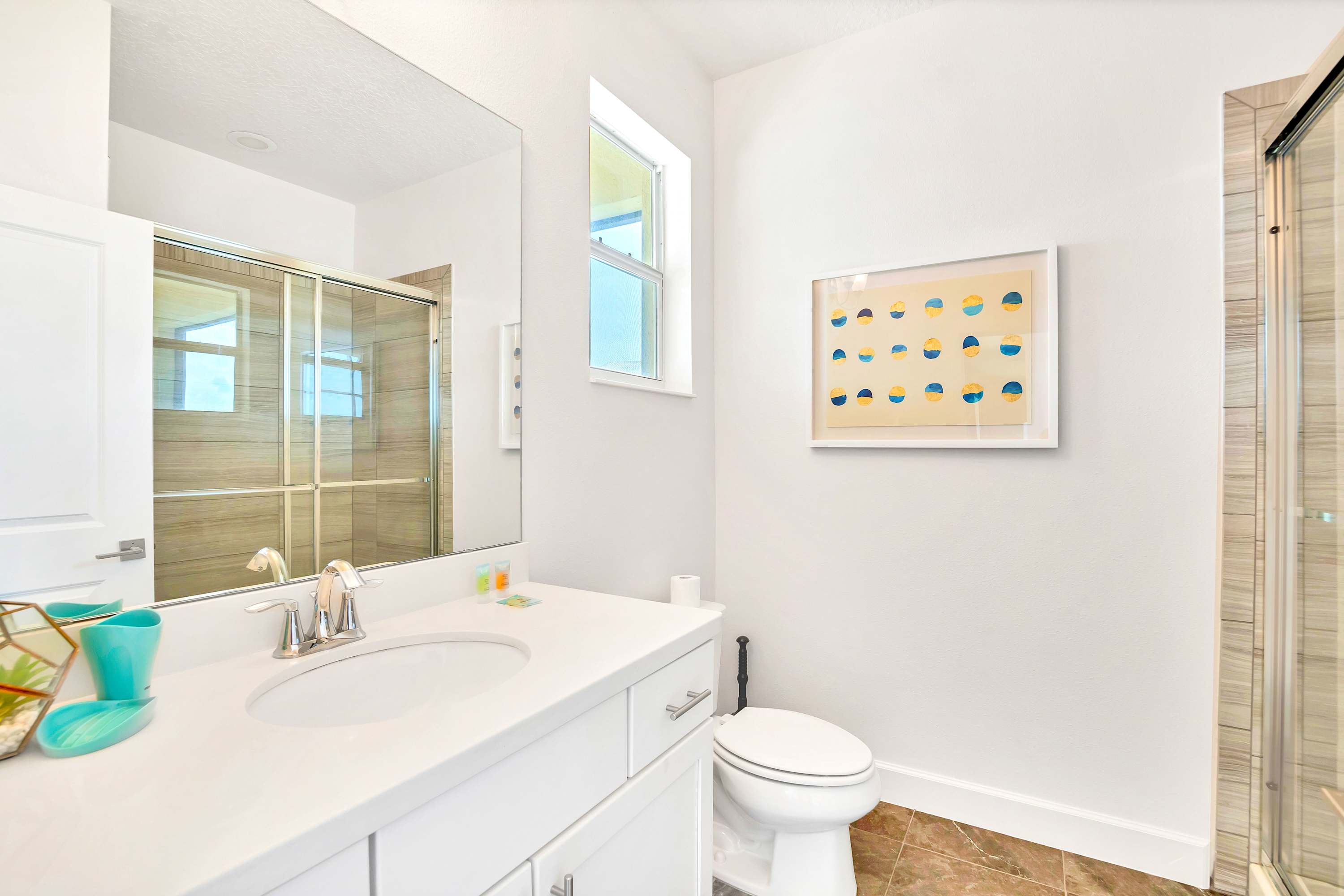 A modern bathroom with a white vanity featuring a single sink and chrome faucet, a large mirror, and a toilet. A glass-enclosed shower with tiled walls is visible, and a window allows natural light, complemented by abstract artwork on the wall.