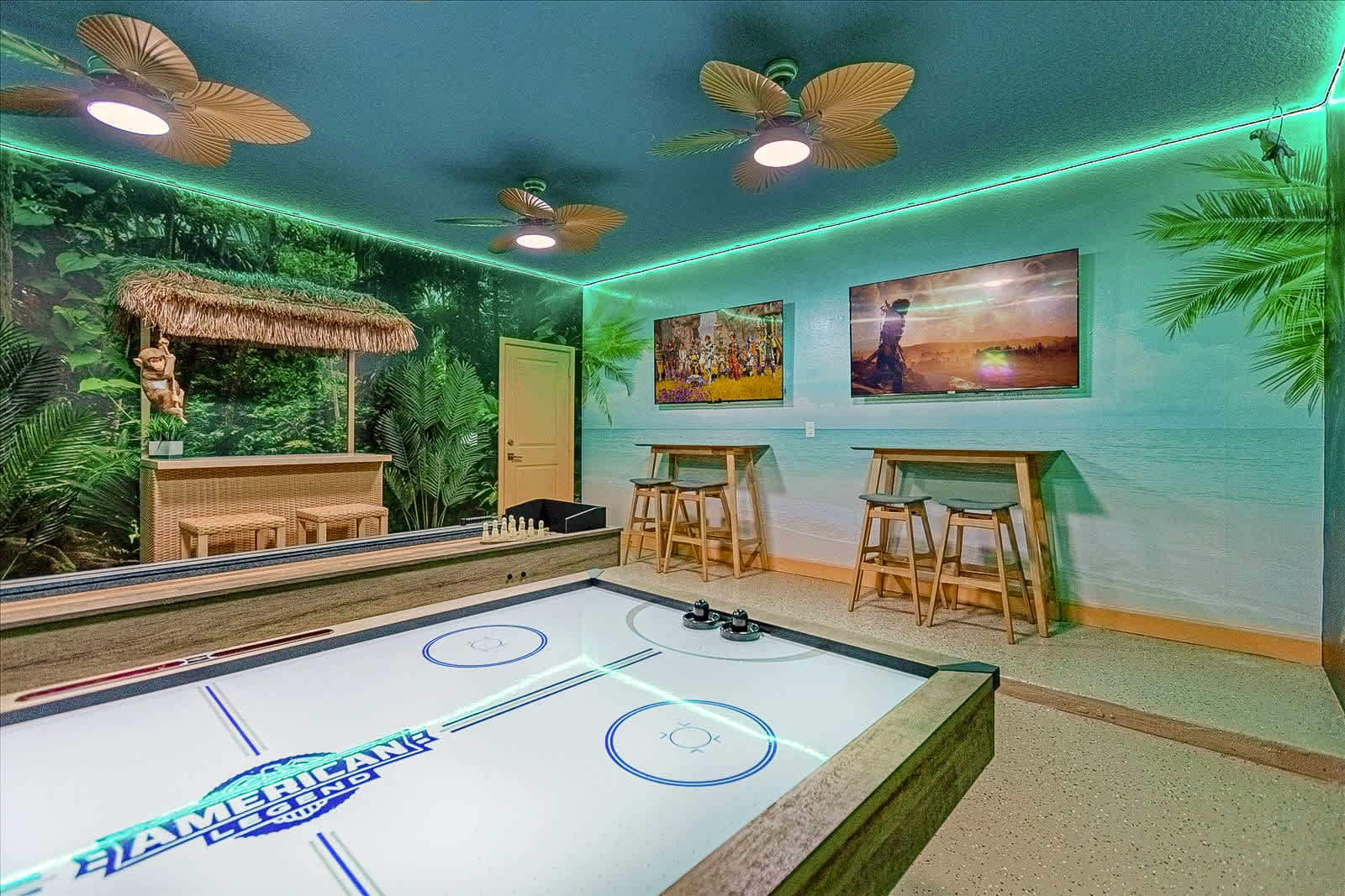 This lively jungle-themed game room is equipped with an air hockey table, a shuffleboard table, and a small tiki bar, all set against a backdrop of artificial plant walls and vibrant murals. Green LED lighting and multiple ceiling fans enhance the playful atmosphere.