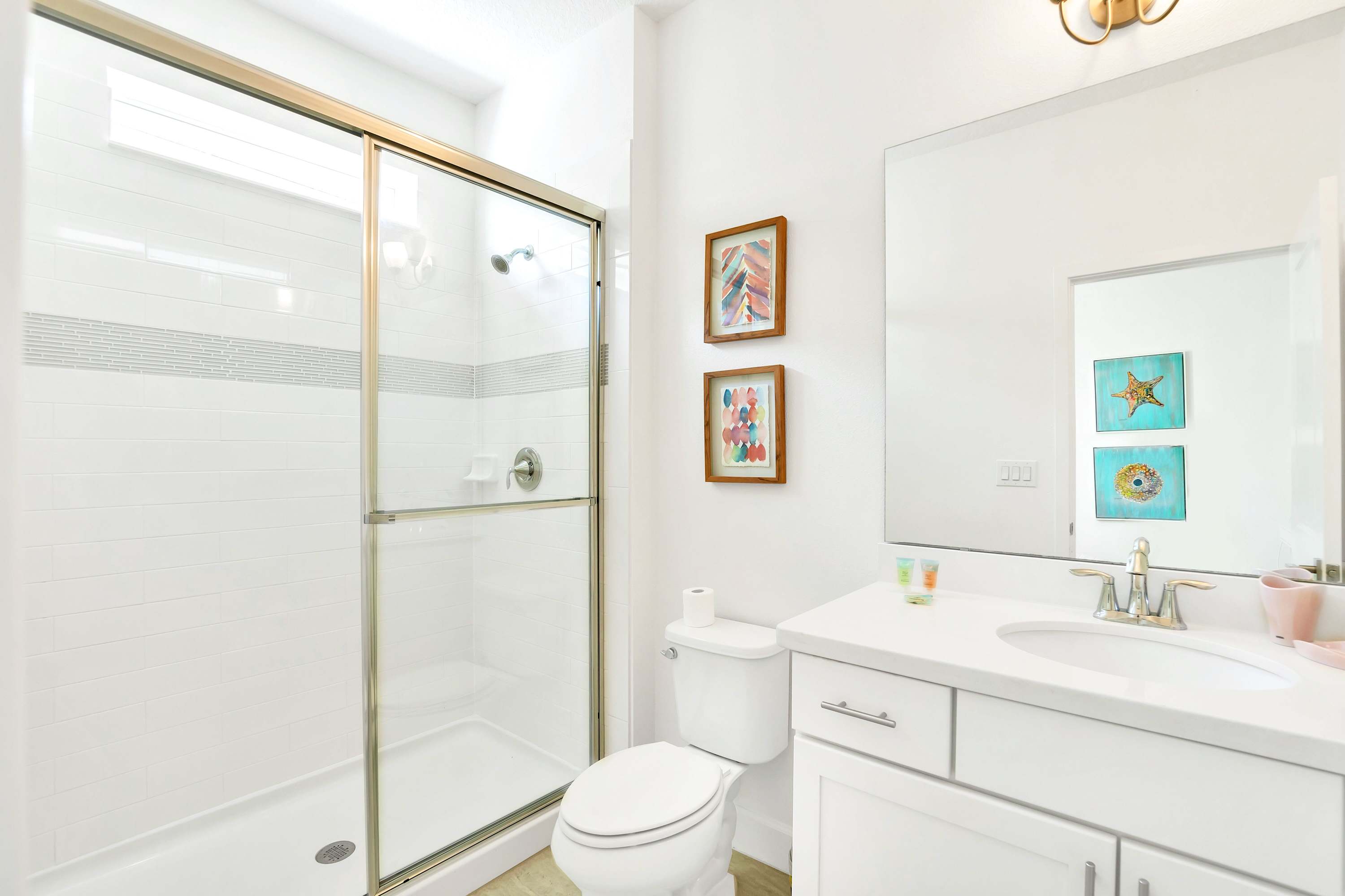Clean white bathroom featuring a single vanity, toilet, and a walk-in shower with a sliding glass door. Decorative framed artwork hangs above the toilet.