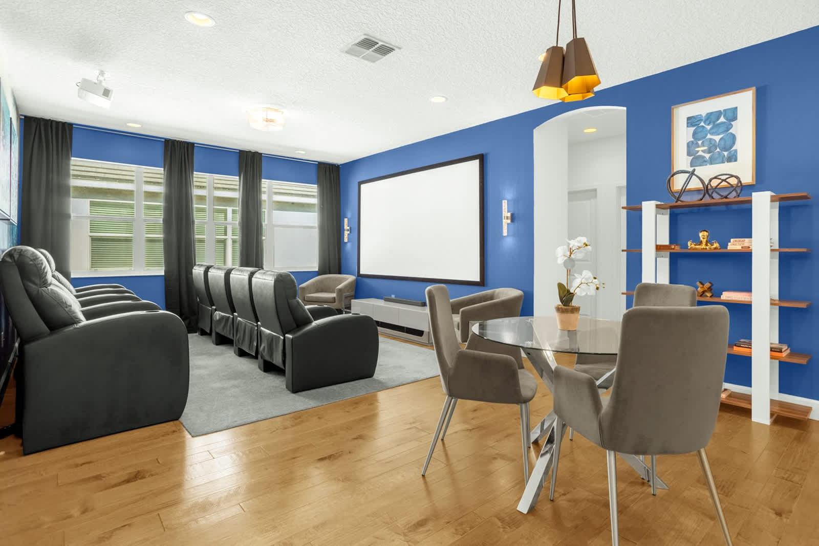 A dedicated media room features a large projection screen, multiple rows of comfortable black reclining theater seats, and a small glass-top dining table with chairs. The room has bright blue accent walls, light wood flooring, and large windows with dark curtains.