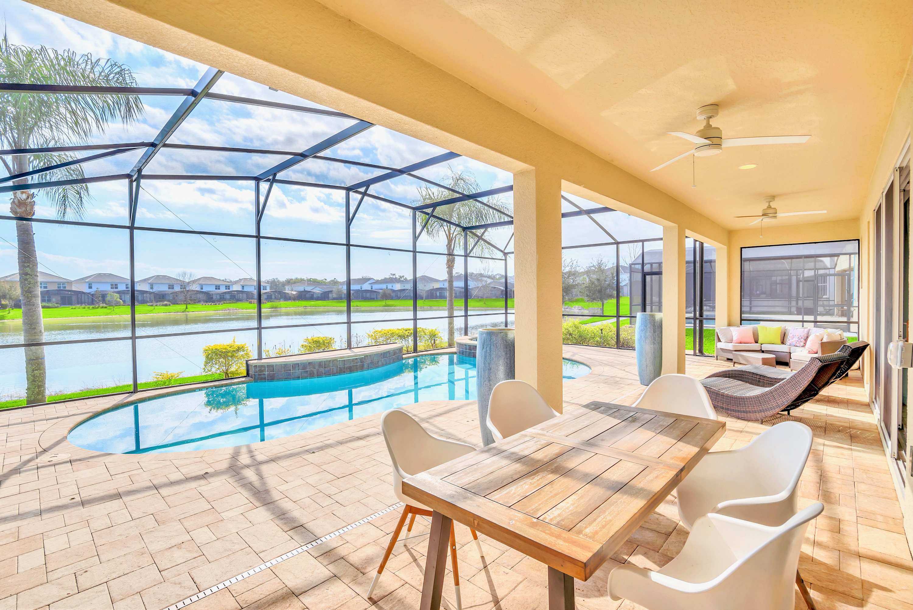A screened-in outdoor pool area features a freeform swimming pool with an integrated hot tub, surrounded by a light-colored paved patio. The space includes an outdoor dining table and lounge seating, all offering expansive views of a lake and distant houses.