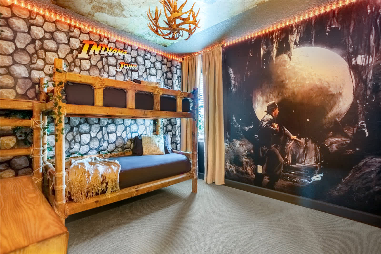 Indiana Jones-themed bedroom featuring a rustic wooden bunk bed, stone-patterned wallpaper, and a large mural depicting a scene from the movie. An antler chandelier hangs from the ceiling.