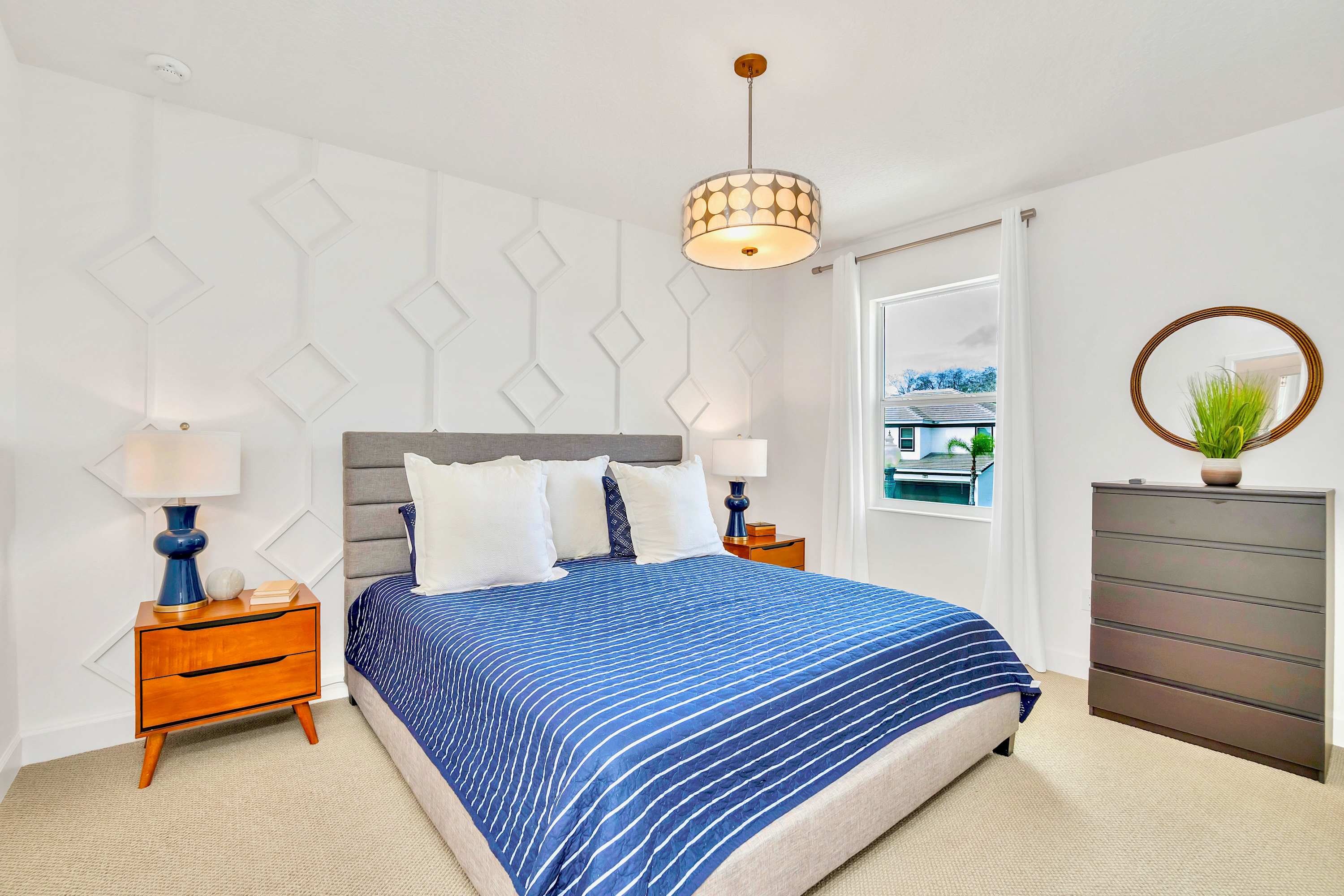 Spacious bedroom featuring a large bed with a gray padded headboard and a blue striped comforter, flanked by wooden nightstands with lamps. A white geometric accent wall adds texture.