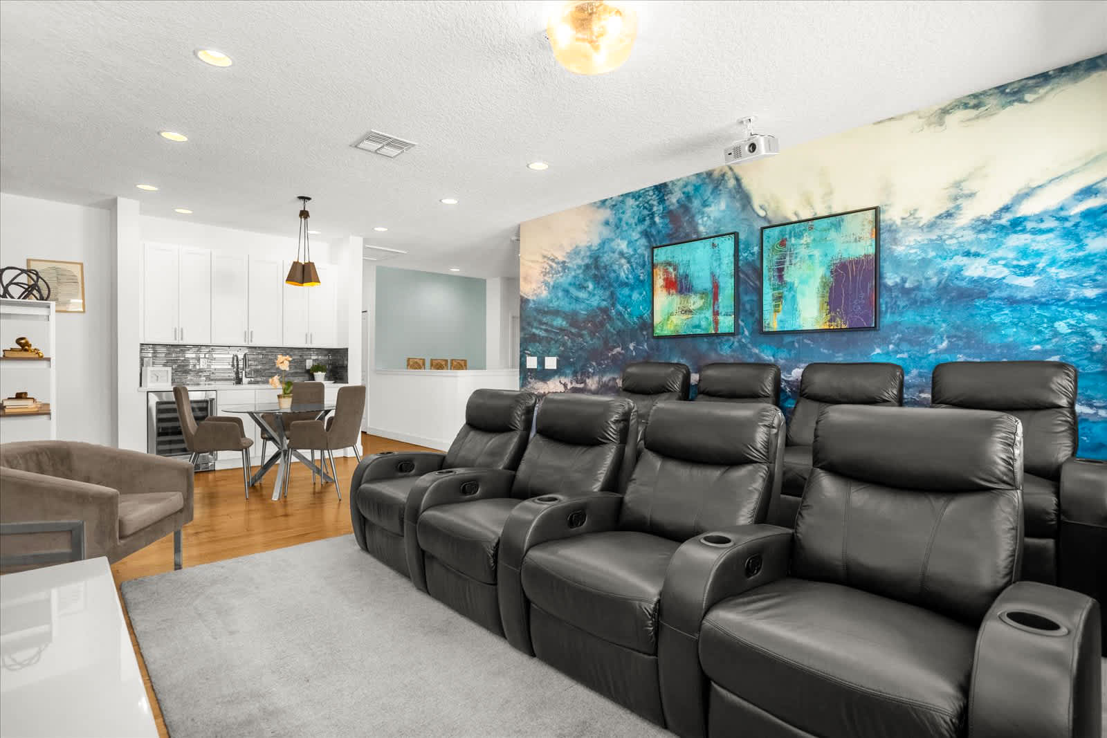 A modern media room features multiple rows of black reclining theater seats facing a large projection screen, with a small kitchenette and dining area visible in the background. The room has a striking blue and white wave-patterned accent wall and light wood flooring.
