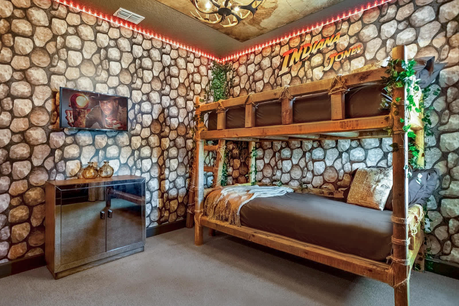 Indiana Jones-themed bedroom with a rustic wooden bunk bed, stone-patterned wallpaper, and an illuminated Indiana Jones logo above the bed. A mirrored cabinet and TV are on the left.