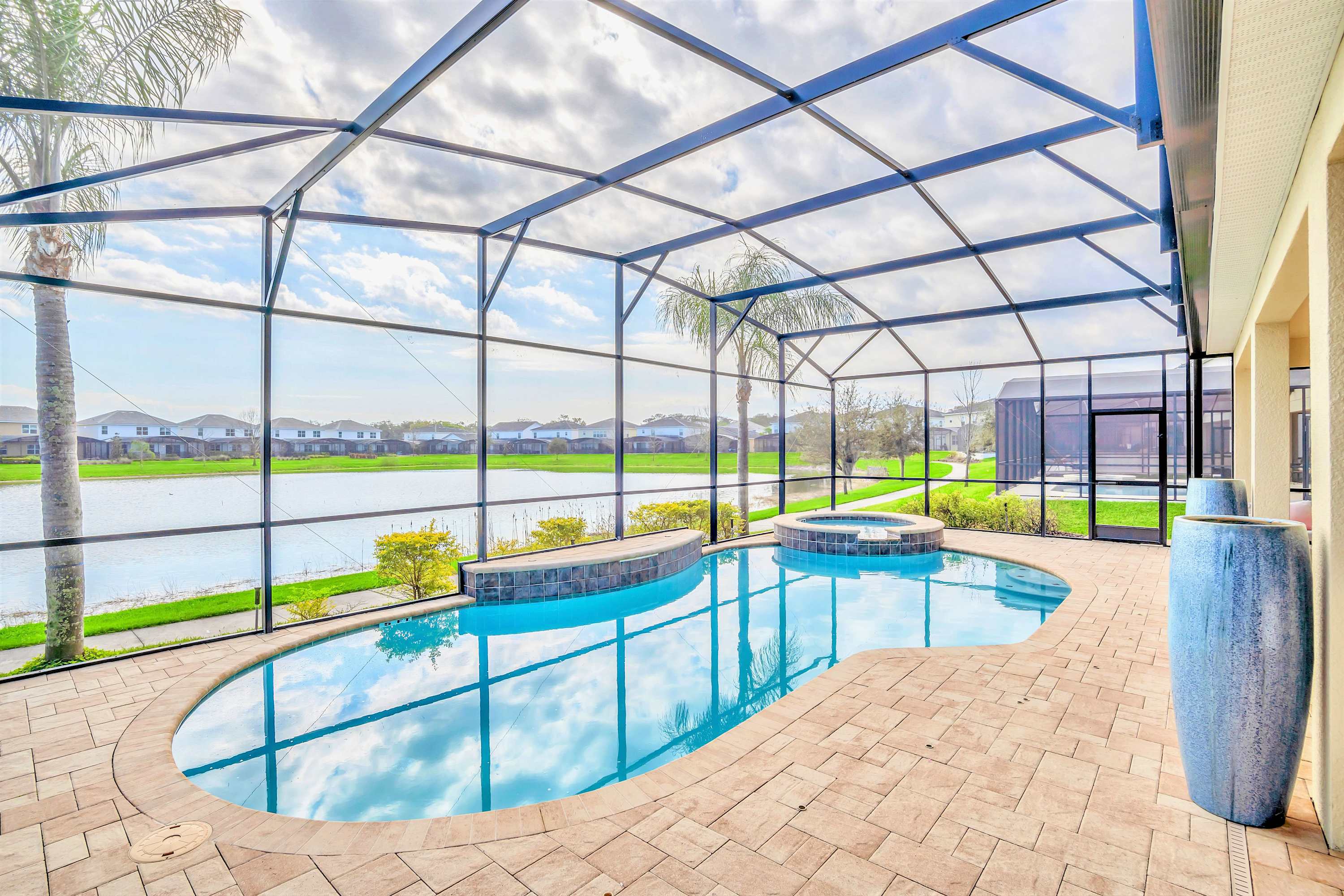 A wide shot of a screened-in private pool and hot tub area, showcasing the light blue water and expansive paved deck. The enclosure provides clear, unobstructed views of a serene lake and residential homes in the background, with a palm tree inside the screen.