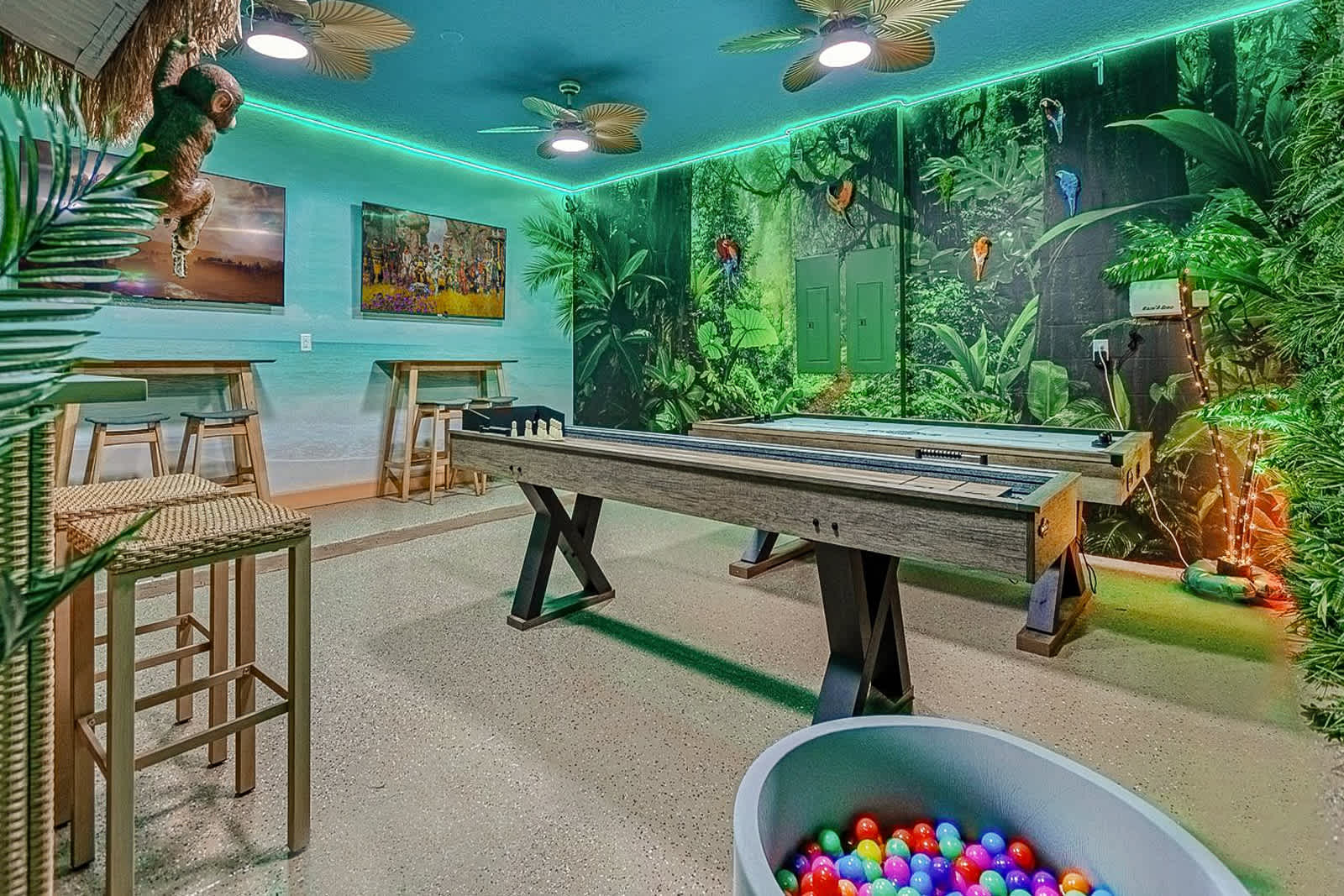 A vibrant jungle-themed game room features a shuffleboard table, a colorful ball pit, and a wall adorned with lush artificial greenery and vibrant animal murals. The room is lit by green LED strips and ceiling fans, with a small bar area in the corner.