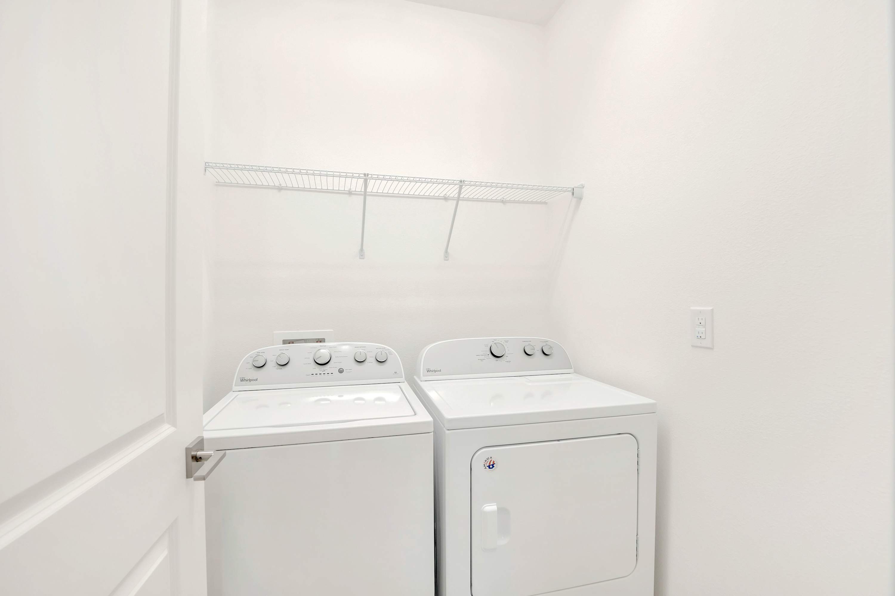 A clean and compact laundry area featuring a white top-loading washing machine and a matching dryer, with a simple wire shelf mounted above. The space is minimalist with white walls, providing essential amenities for guests.
