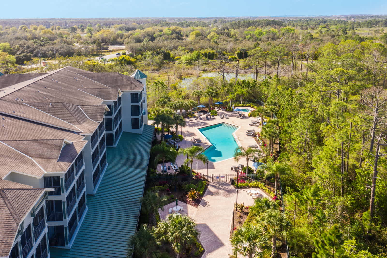 An aerial view showcasing the condominium building alongside a large communal swimming pool with lounge chairs, surrounded by abundant green trees and natural landscape.