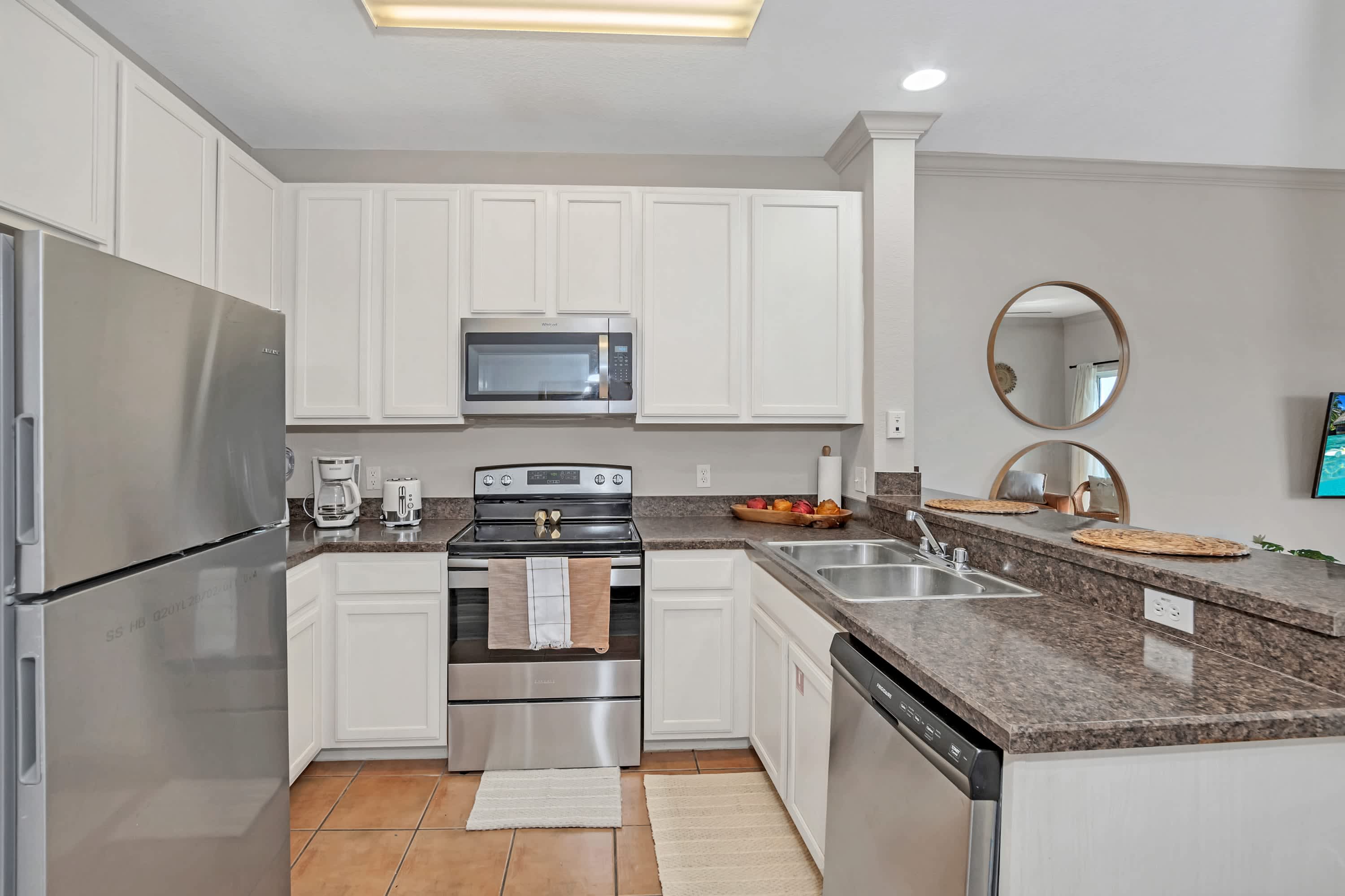 A modern kitchen with white shaker cabinets, dark speckled countertops, stainless steel appliances including a refrigerator, oven, microwave, and dishwasher, and a double sink. The floor is tiled in a terracotta color.