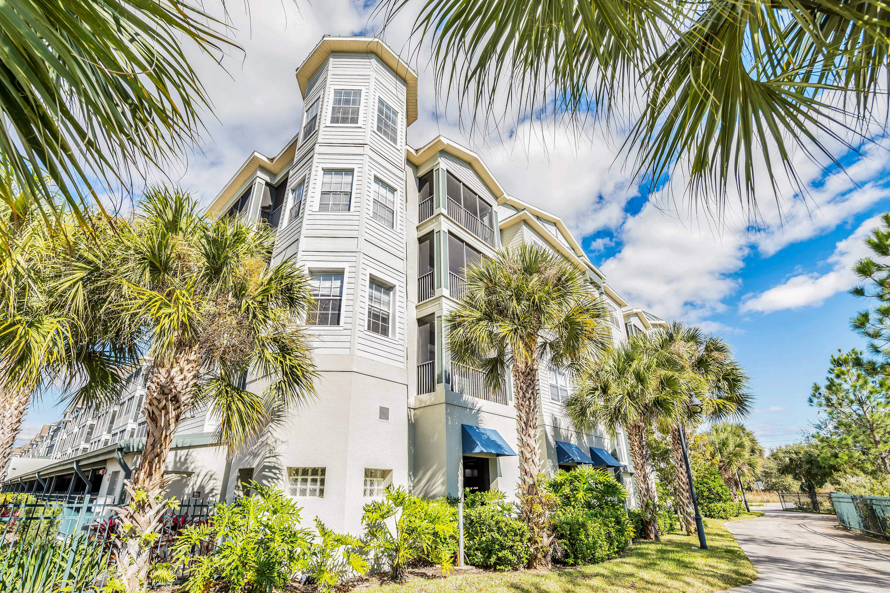A multi-story condominium building with light-colored siding, bay windows, and screened balconies is framed by lush palm fronds, set against a partly cloudy blue sky.