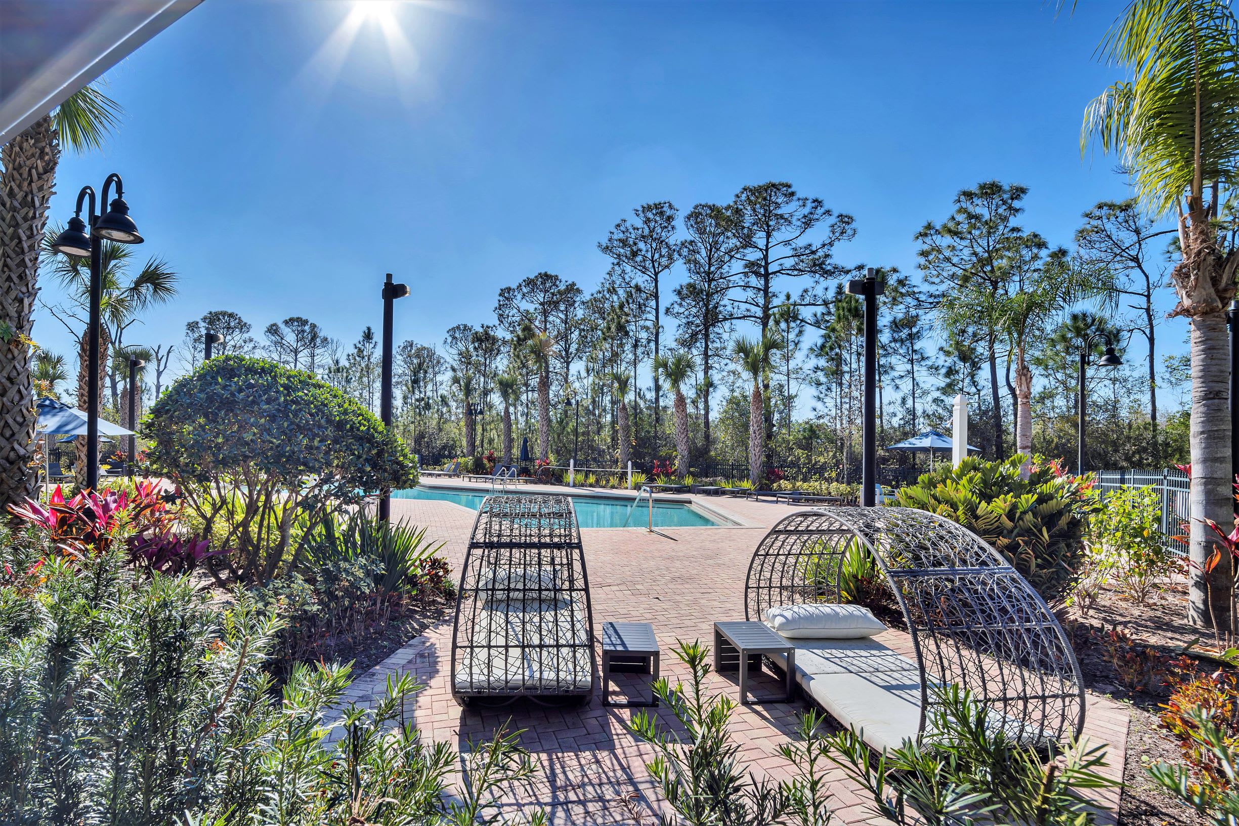A sunny outdoor pool area featuring modern woven lounge chairs and small side tables on a red brick paver deck. Lush green landscaping and tall trees surround the pool under a clear blue sky.