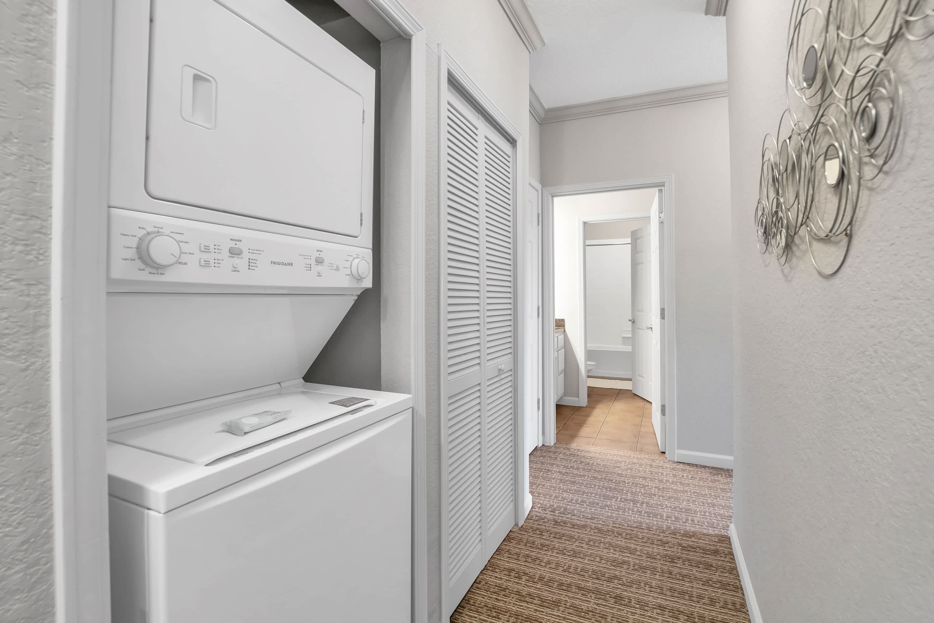 A compact laundry area located in a hallway closet, featuring a stacked white washer and dryer unit. The hallway has carpeted flooring and leads to other rooms.