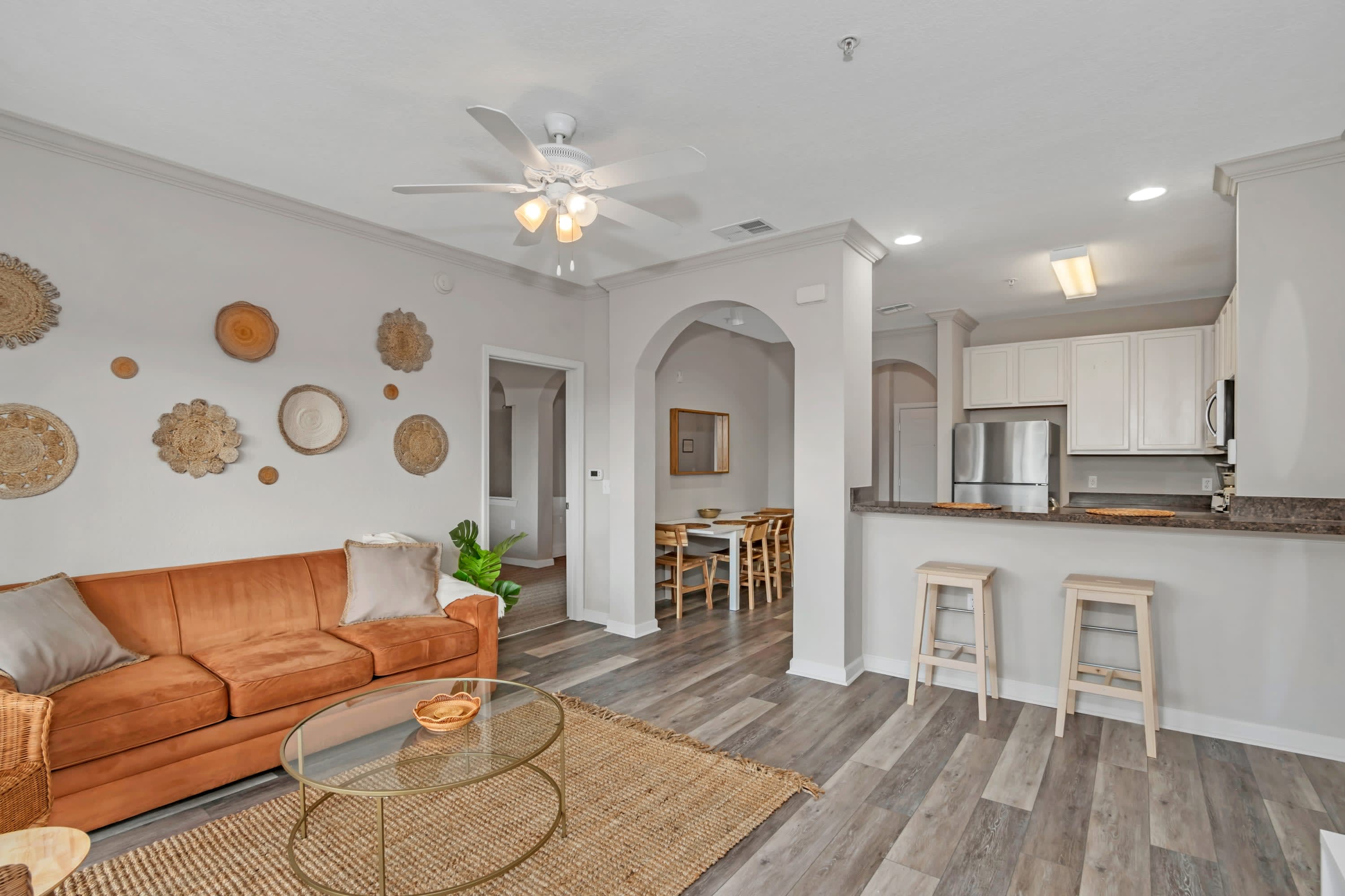 An open-concept living area with an orange sofa and a view towards the kitchen and dining area through arched doorways. The space features wood-look flooring and decorative woven wall art.