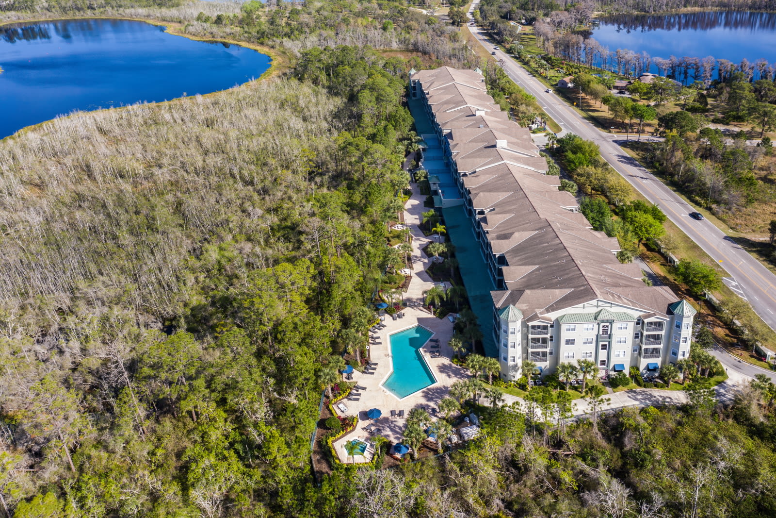 An aerial view showcases the long, multi-story condominium building with a large outdoor swimming pool and lounge area, nestled between dense natural foliage and two serene lakes, with a road running parallel to the property.
